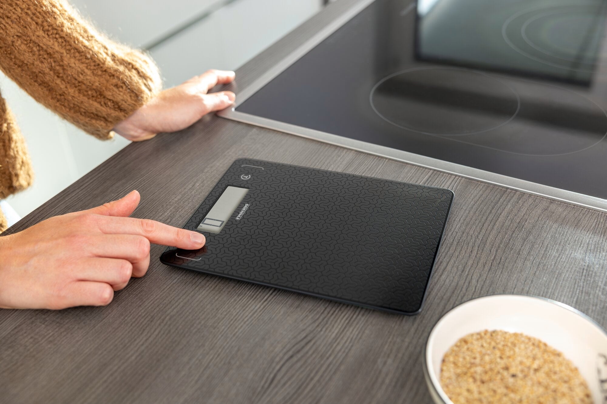 A person is using the Comfort Mosaic digital kitchen scale on a kitchen worktop. The scale has a modern design and an easy-to-read display. Next to it is a bowl of grains.