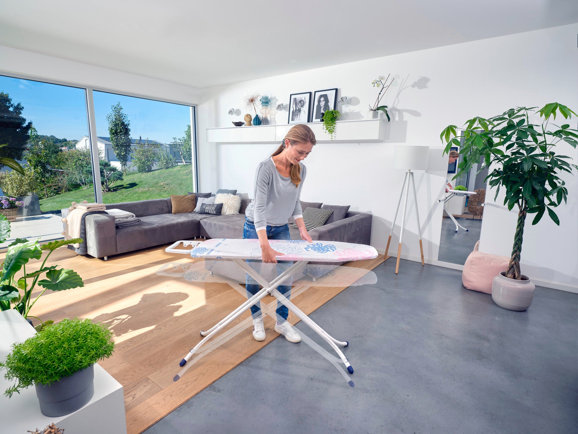 Eine Frau bügelt auf dem Bügeltisch Classic M Compact in einem modernen Wohnzimmer mit großen Fenstern und Pflanzen. Der Tisch ist stabil und bietet eine großzügige Bügelfläche.