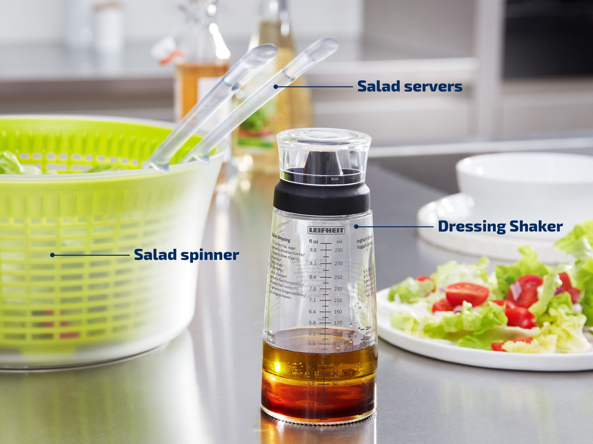 Auf einer modernen Küchenarbeitsplatte stehen ein grüner Salatschleuder, ein Dressing-Shaker und Salatbesteck. Im Hintergrund ist ein frischer Salat mit Tomaten zu sehen.
