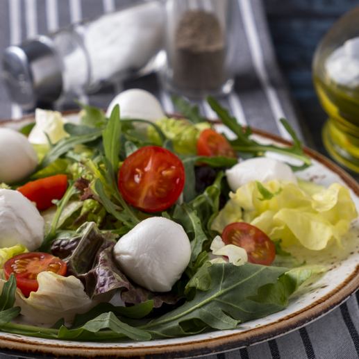 Ein Teller mit frischem Salat aus Rucola, Kopfsalat, Kirschtomaten und kleinen Mozzarellakugeln, serviert auf einer gestreiften Tischdecke.