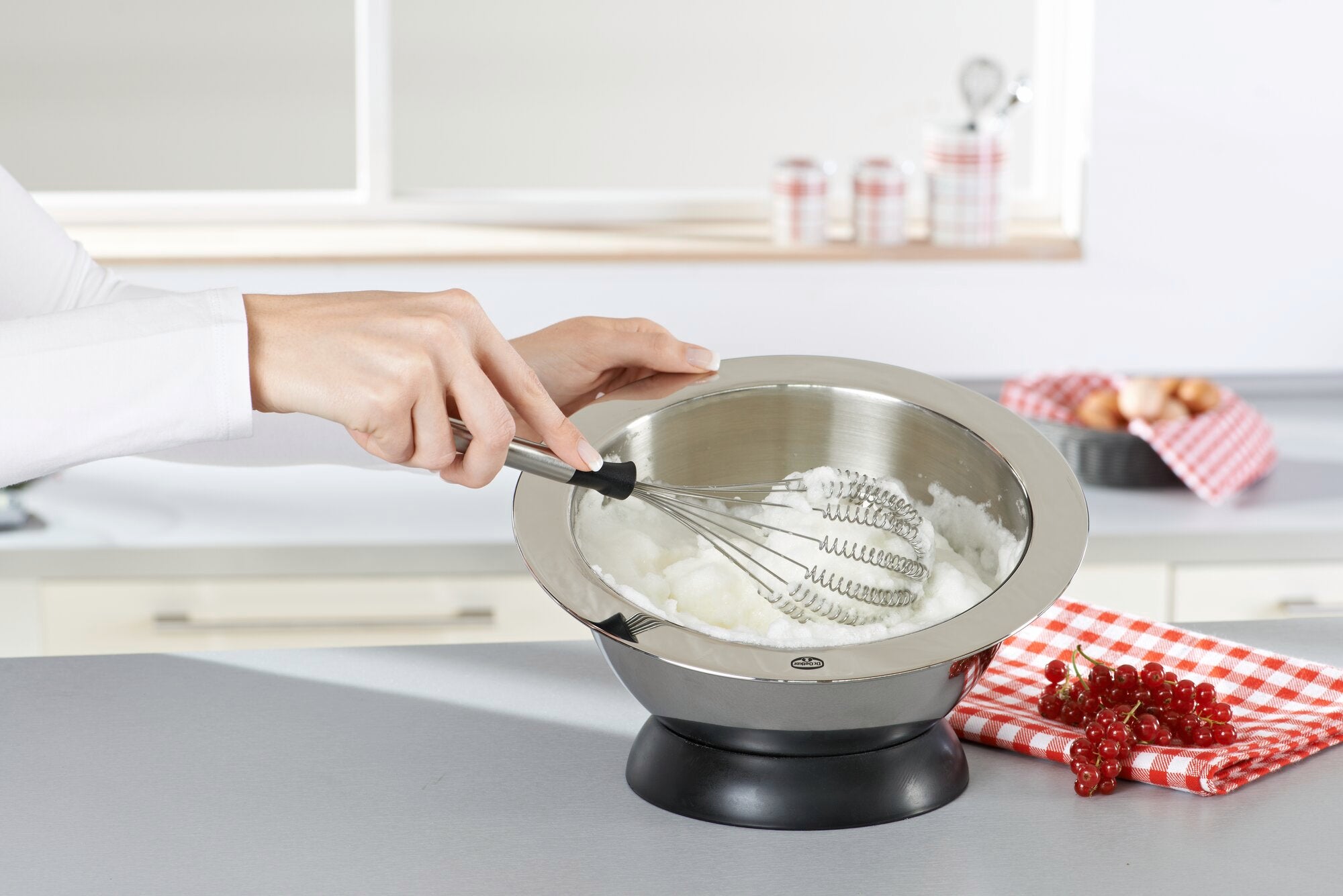 Una persona utilizza la frusta Speed Quirl S per mescolare gli ingredienti in una ciotola di metallo in una cucina moderna. Sullo sfondo si vedono utensili da cucina e decorazioni.