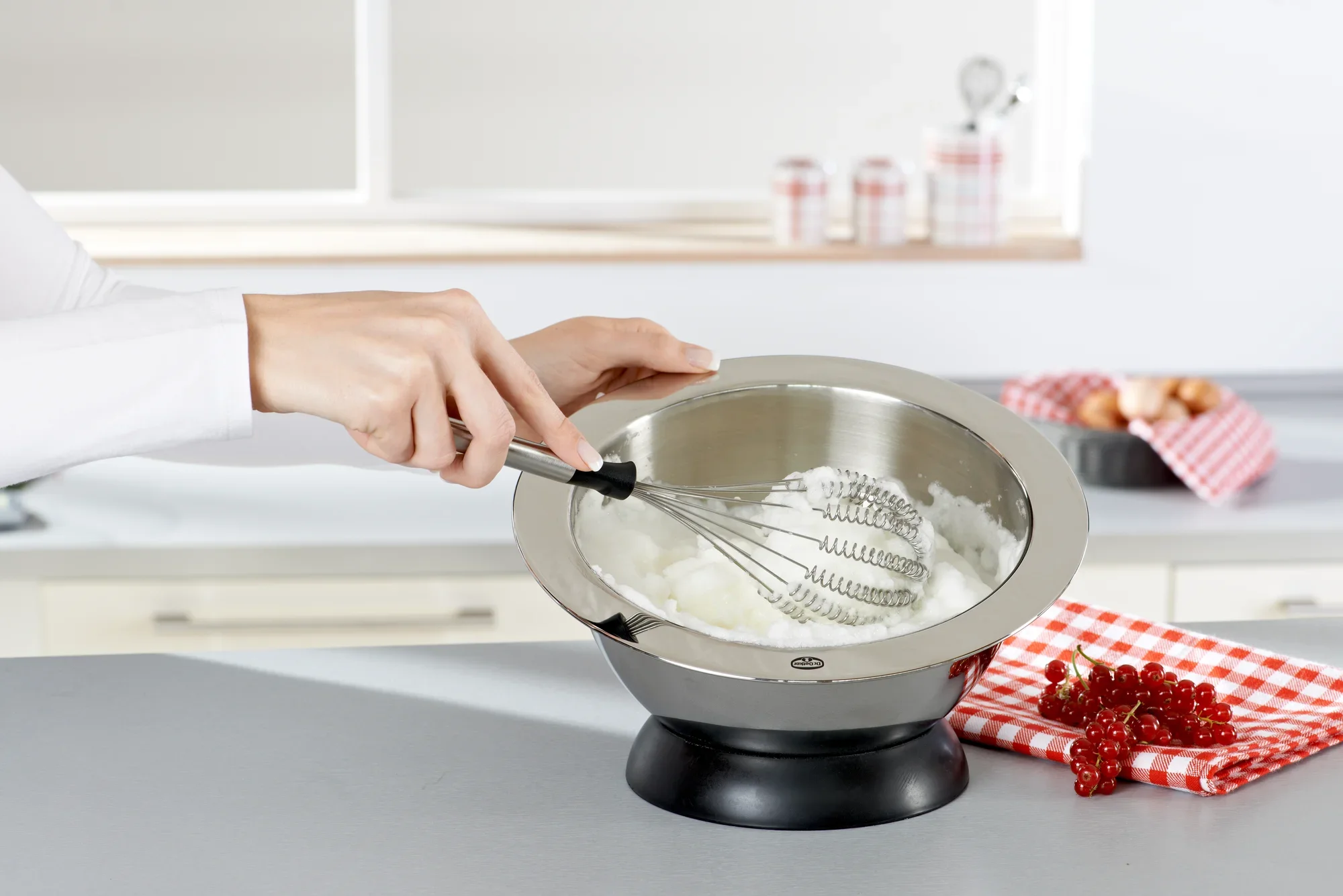 Une personne utilise le fouet Speed Quirl S pour mélanger des ingrédients dans un saladier en métal dans une cuisine moderne. On aperçoit des ustensiles de cuisine et des décorations en arrière-plan.