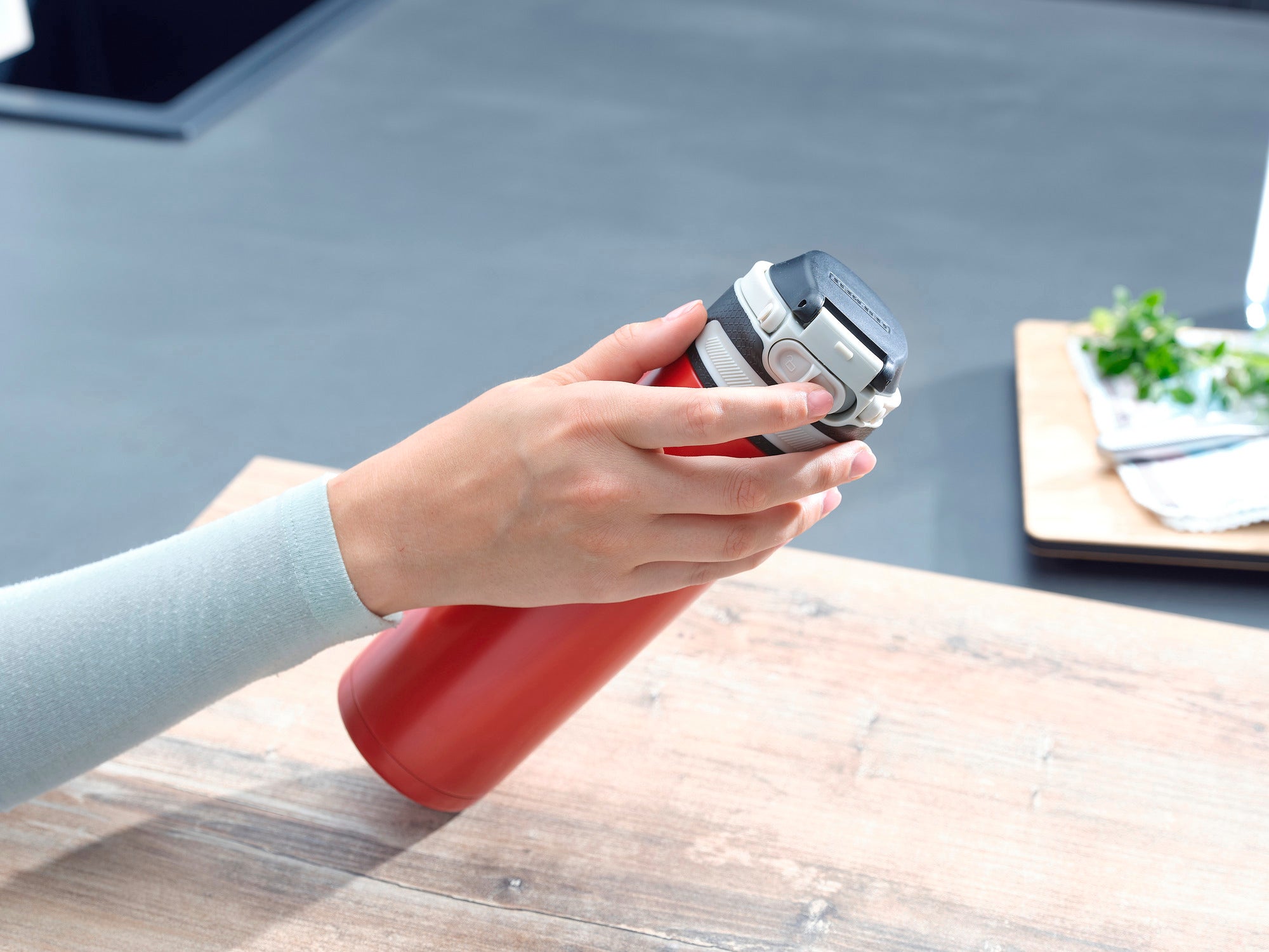 Tasse isotherme Flip 600 ml en rouge sur une table. Une main ouvre le couvercle. Des ustensiles de cuisine sont visibles à l'arrière-plan. Idéale pour les déplacements, elle garde les boissons au chaud.