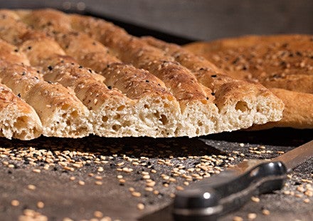 Frisches, aufgeschnittenes Fladenbrot mit Sesam und Schwarzkuemmel liegt auf einem Backblech neben verstreutem Sesam und einem Messer.