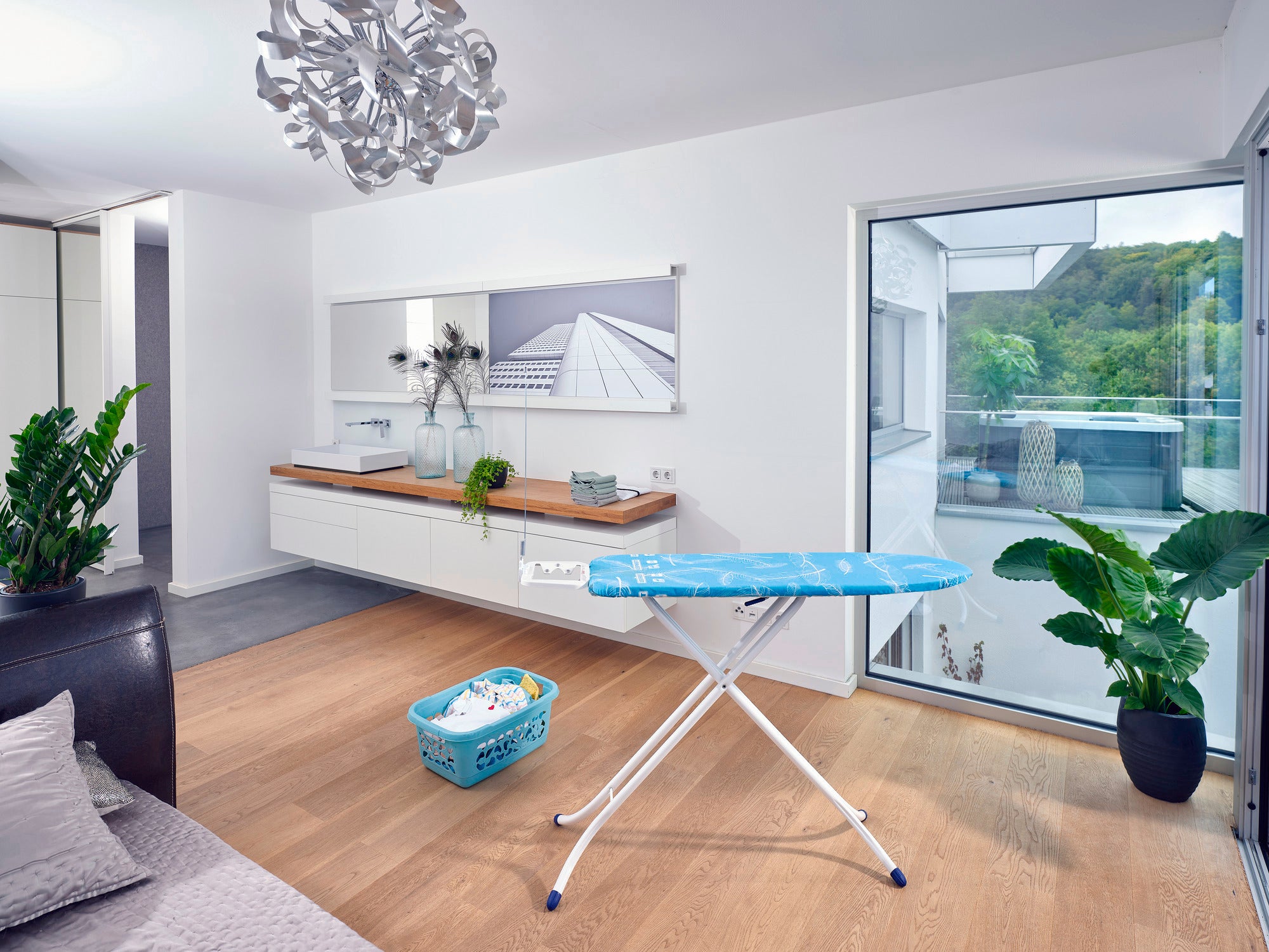 Air Board M Compact ironing board in a modern, bright room with large windows. The board stands on a wooden floor, surrounded by plants and a laundry basket.