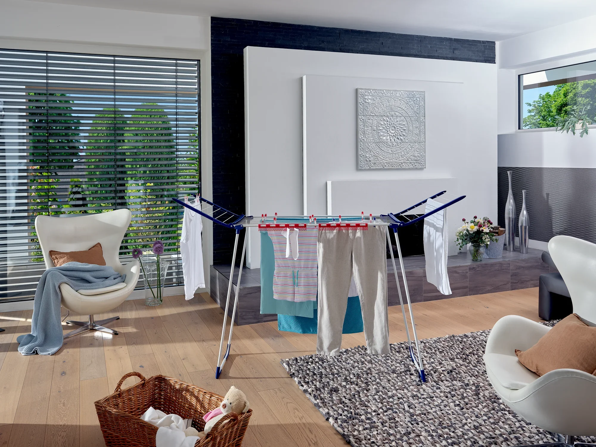 Ein Standtrockner Pegasus 180 MAXX steht in einem modernen Wohnzimmer. Er ist mit verschiedenen Kleidungsstücken behängt. Im Hintergrund sind stilvolle Möbel und Dekorationen zu sehen.