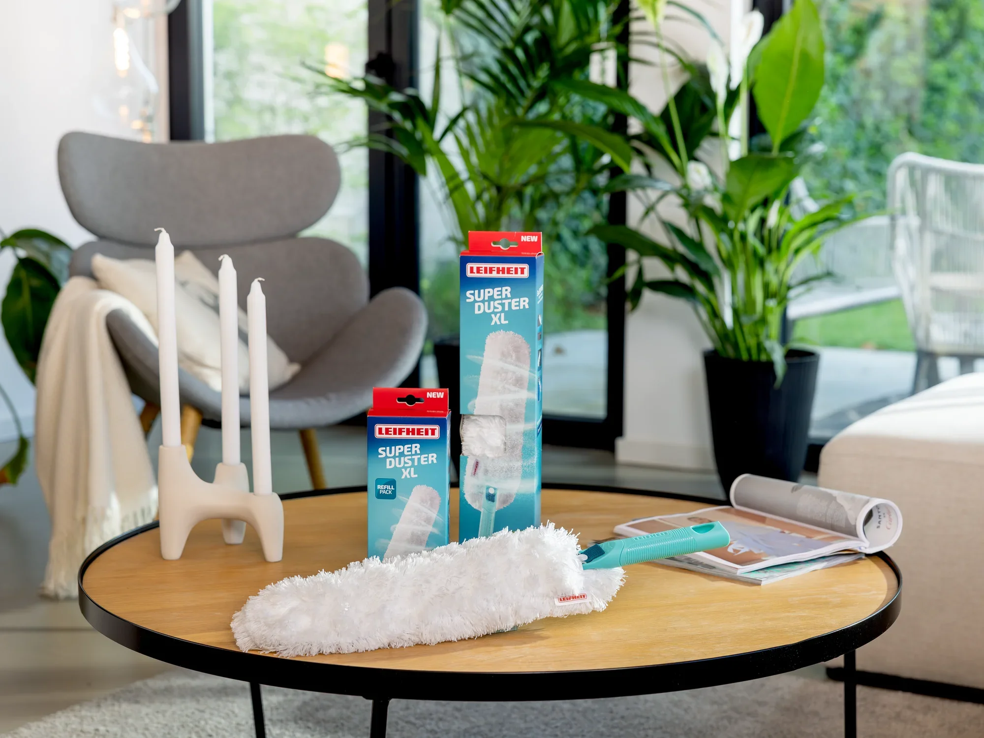 Una sala de estar moderna con una mesa redonda de madera sobre la que se encuentra un plumero SUPERDUSTER XL. Al fondo se ven plantas, un sillón y unos candelabros. Ideal para eliminar el polvo de forma eficaz.