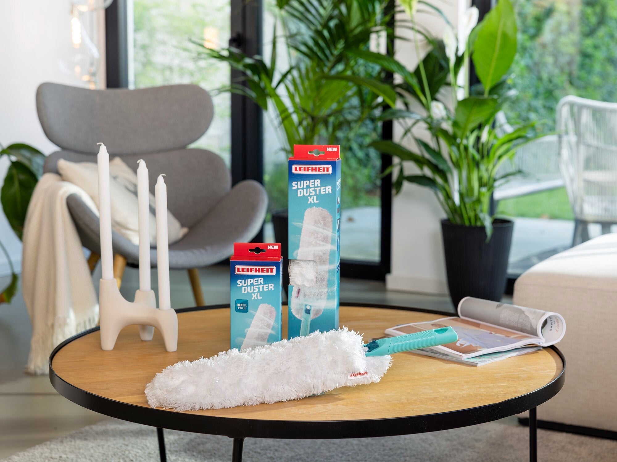 Une pièce moderne avec une table ronde en bois sur laquelle repose un plumeau SUPERDUSTER XL. À l'arrière-plan, on aperçoit des plantes, un fauteuil et des bougeoirs. Idéal pour éliminer efficacement la poussière.