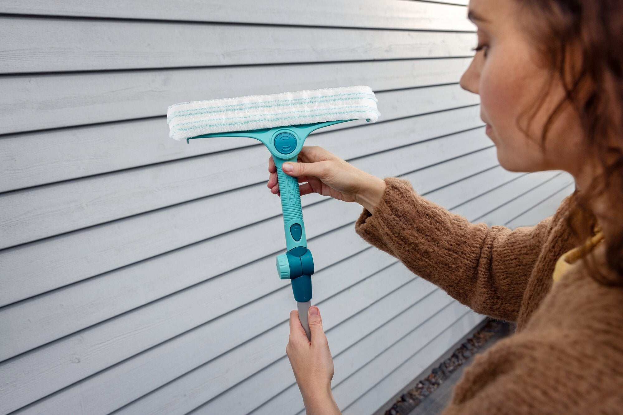 Eine Person hält den Window and Frame Cleaner 4in1 mit Teleskopstange vor einer Holzverkleidung. Der Reiniger hat einen blauen Griff und einen weißen Reinigungskopf.