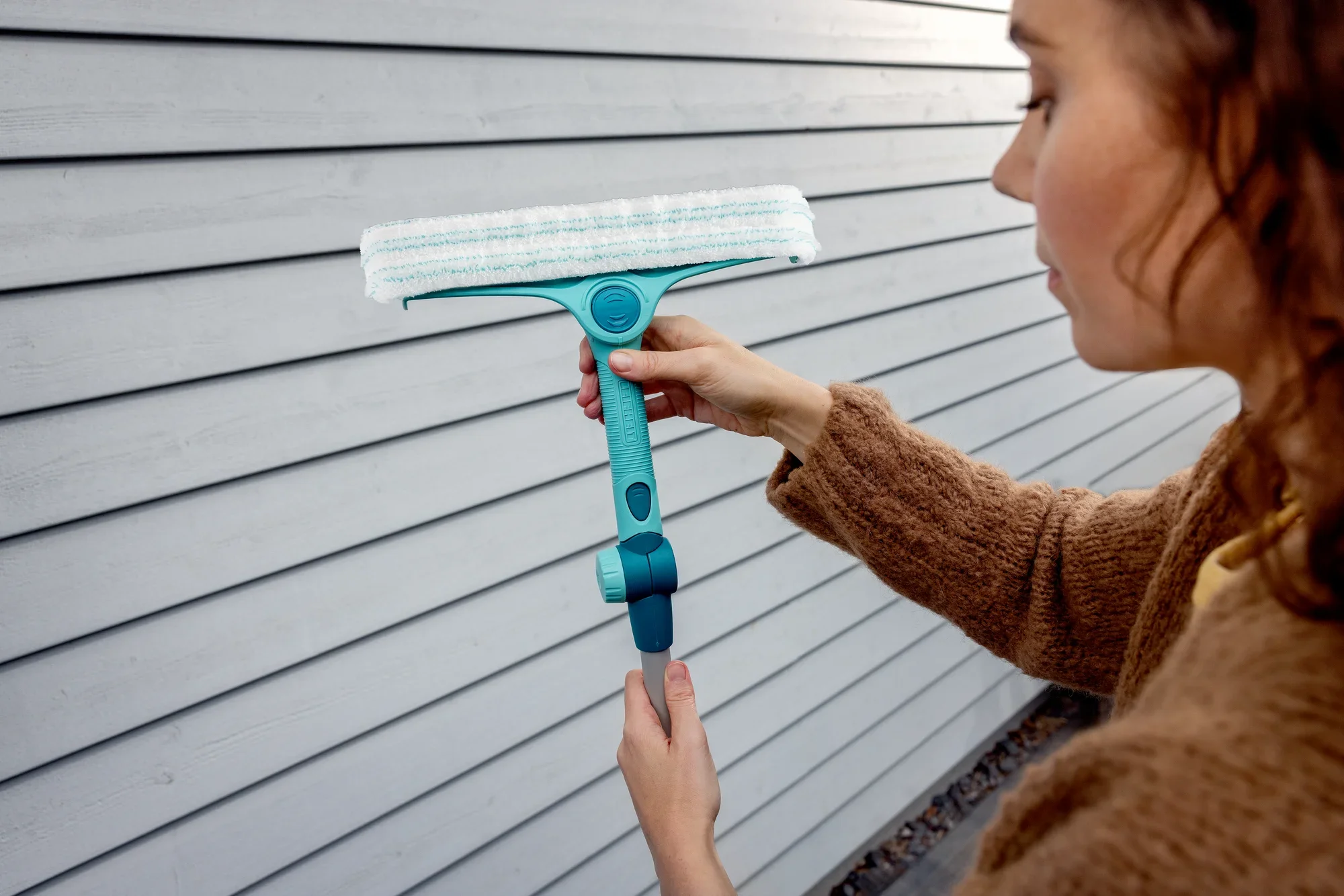 Een persoon houdt de Raam- en kozijnreiniger 4in1 met telescoopstang voor een houten lambrisering. De reiniger heeft een blauwe steel en een witte reinigingskop.