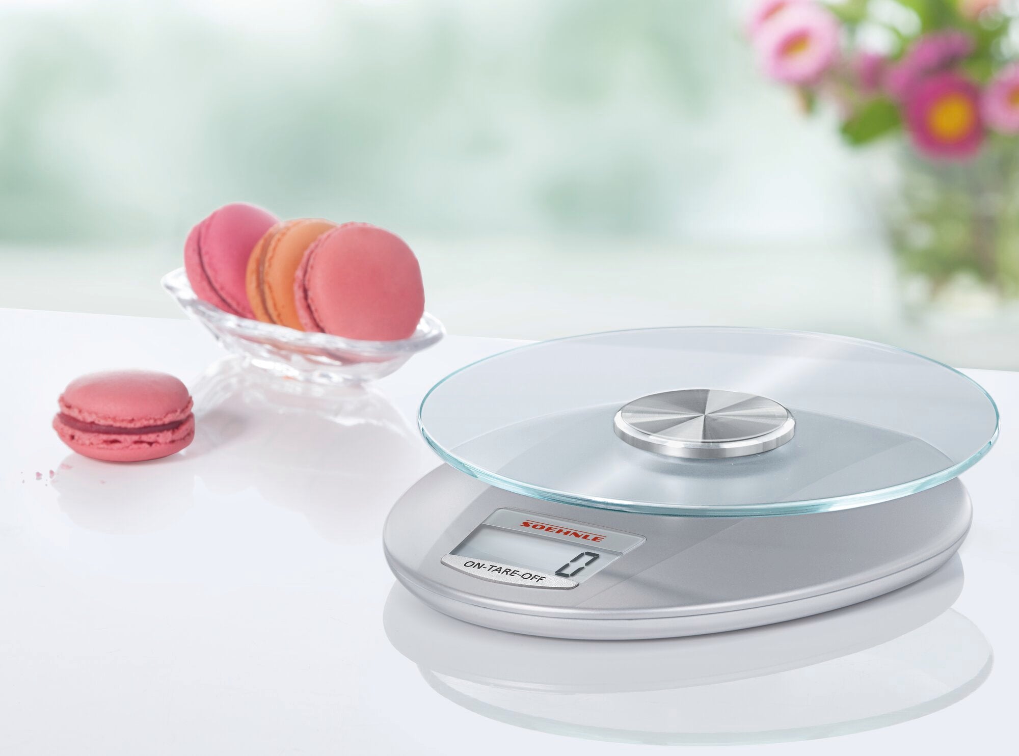 Eine digitale Küchenwaage in Silber, Modell Roma, steht auf einer weißen Oberfläche. Daneben liegt eine Schale mit rosa Macarons. Die Waage hat ein modernes Design mit Glasplatte.