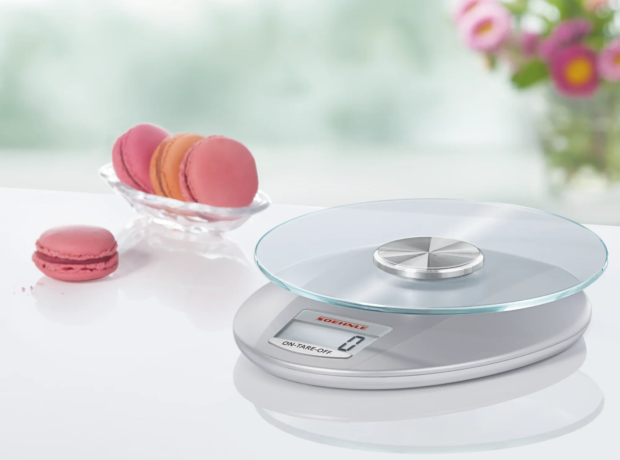 Cyfrowa waga kuchenna w kolorze srebrnym, model Roma, stoi na białej powierzchni. Obok stoi miska z różowymi makaronikami. Waga ma nowoczesny design ze szklaną płytą.