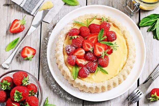 Eine Erdbeertarte mit gelber Creme, dekoriert mit frischen Erdbeeren und Minzblaettern, auf einem Holztisch mit Besteck und Erdbeeren.