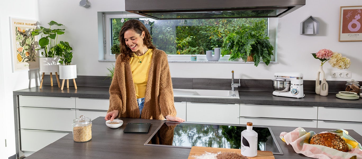 Frühlingsrezepte entdecken: gesund und lecker Eine Frau steht in einer modernen Kueche und wiegt mit einer digitalen Kuechenwaage von Leifheit Zutaten fuer ihr Muesli ab.
