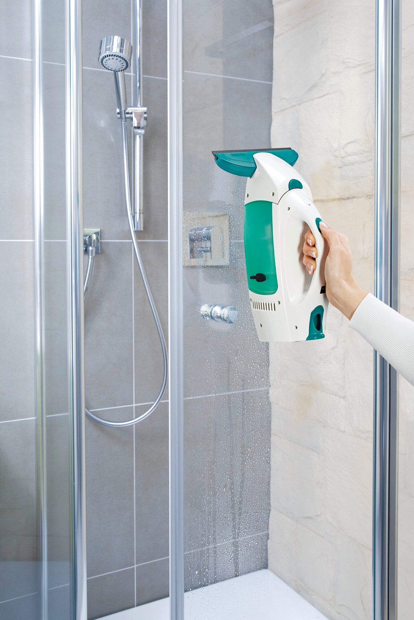 Een Dry and Clean raamstofzuiger met een kleine zuigmond wordt gebruikt in een douche. Eén hand houdt het apparaat vast terwijl het water uit de douchewand zuigt. Ideaal voor streeploos schoonmaken.