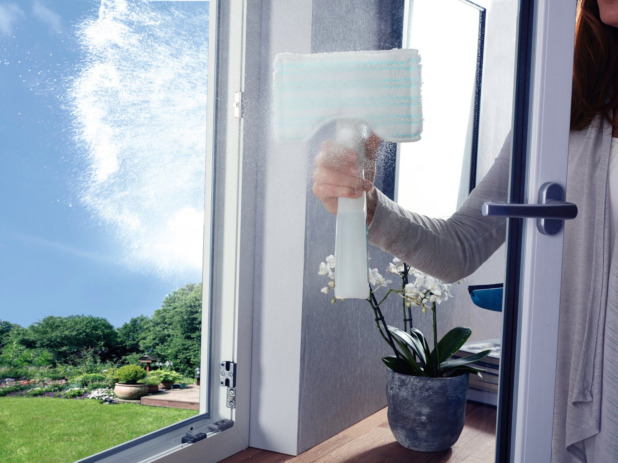 Fenster-Sprühwischer im Einsatz: Eine Person reinigt ein geöffnetes Fenster mit dem Fenster-Sprühwischer. Im Hintergrund ist ein Garten mit Pflanzen zu sehen.