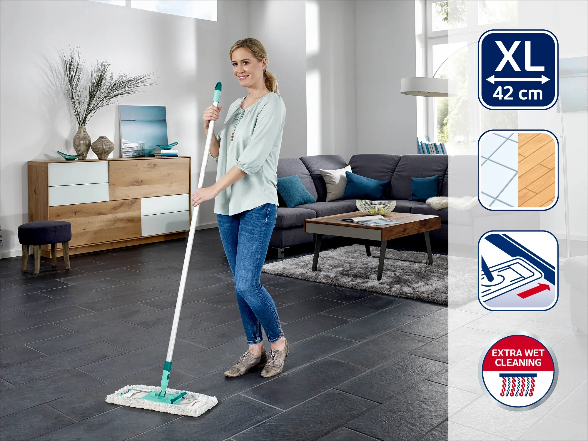 Een vrouw maakt de vloer van een moderne woonkamer schoon met de Leifheit dweilmop. De kamer is stijlvol ingericht, met een grote bank en een dressoir. De mop heeft een XL-breedte van 42 cm voor efficiënt schoonmaken.
