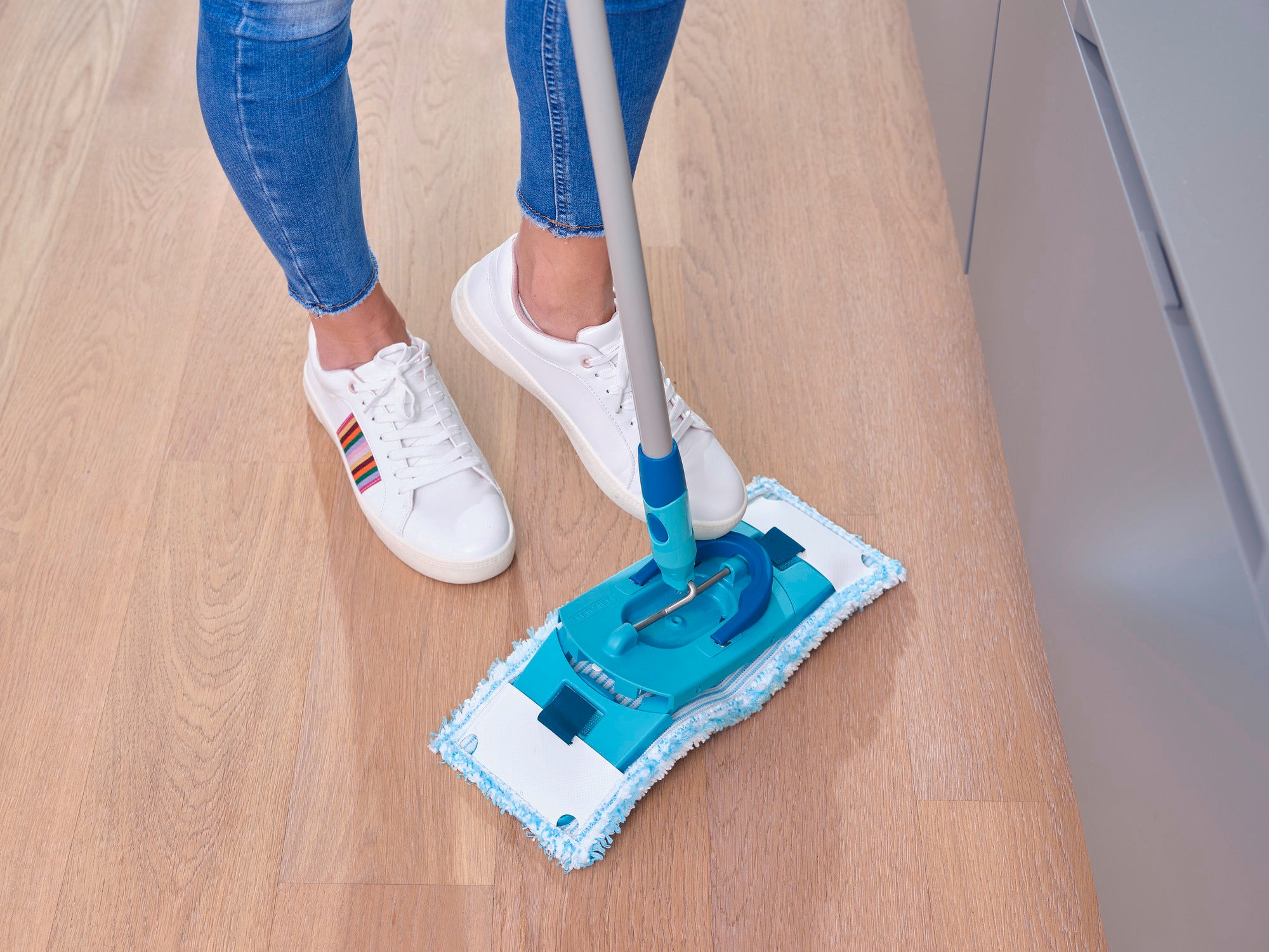 Eine Person reinigt einen Holzboden mit dem CLEAN TWIST M Ergo. Der Wischmopp hat ein blaues Design und wird von einer Person in Jeans und weißen Sneakers verwendet. Ideal für gründliche Reinigung.