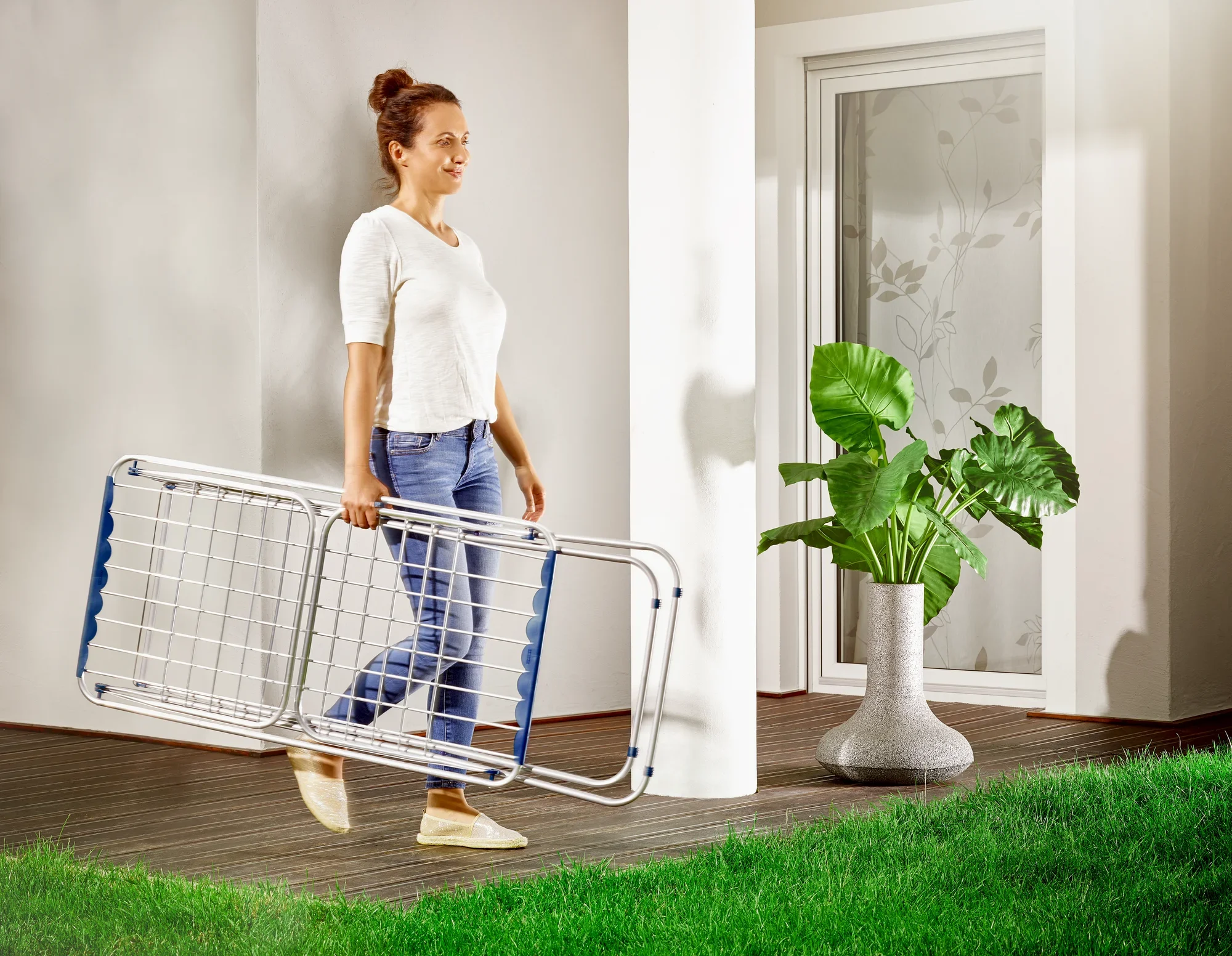 Een vrouw draagt de Siena 180 aluminium wasdroger op voet door een moderne kamer met een houten vloer. Op de achtergrond staat een grote plant in een elegante pot.
