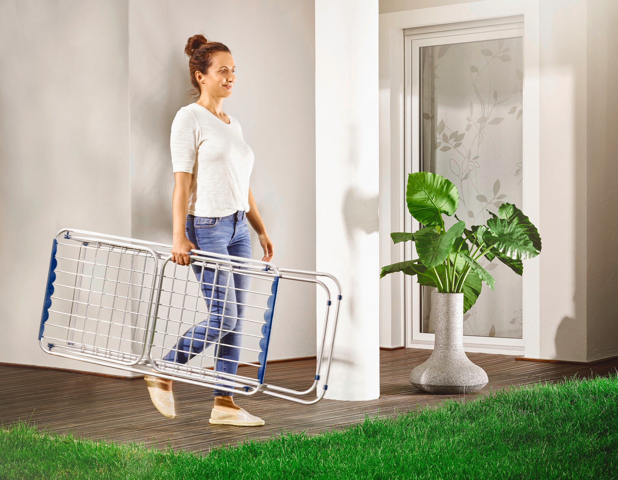 Een vrouw draagt de Siena 180 aluminium wasdroger op voet door een moderne kamer met een houten vloer. Op de achtergrond staat een grote plant in een elegante pot.