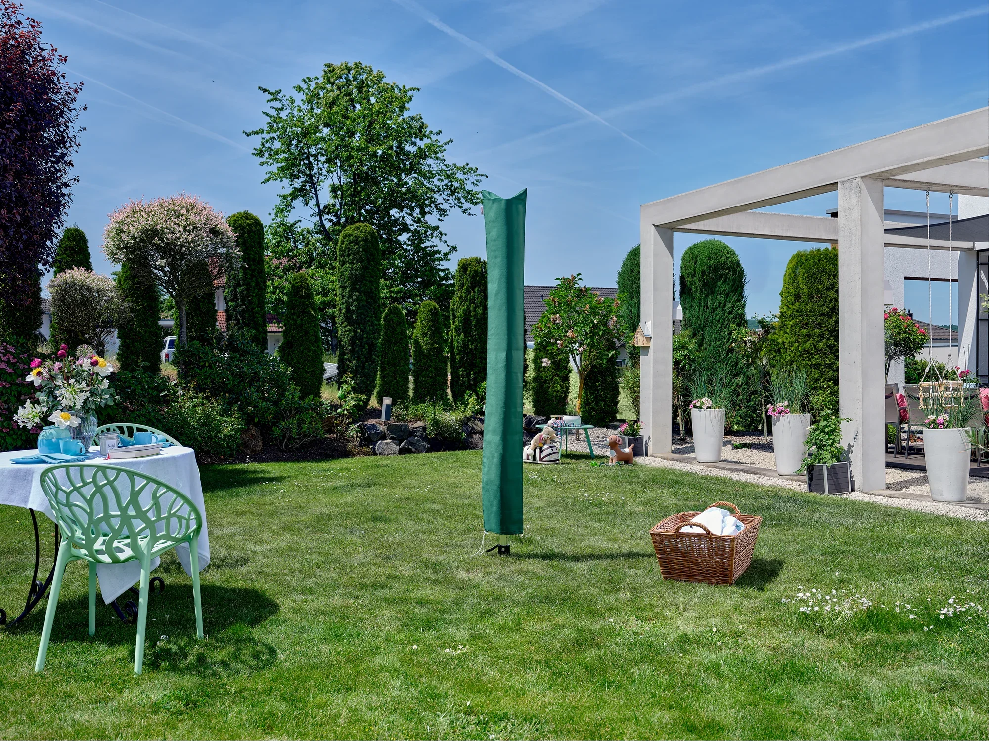 Zielona suszarka obrotowa Leifheit Linomatic 500 Deluxe stoi w zadbanym ogrodzie. W tle widać rośliny i nowoczesny pawilon. Scenę dopełnia stół z kwiatami i kosz na pranie.