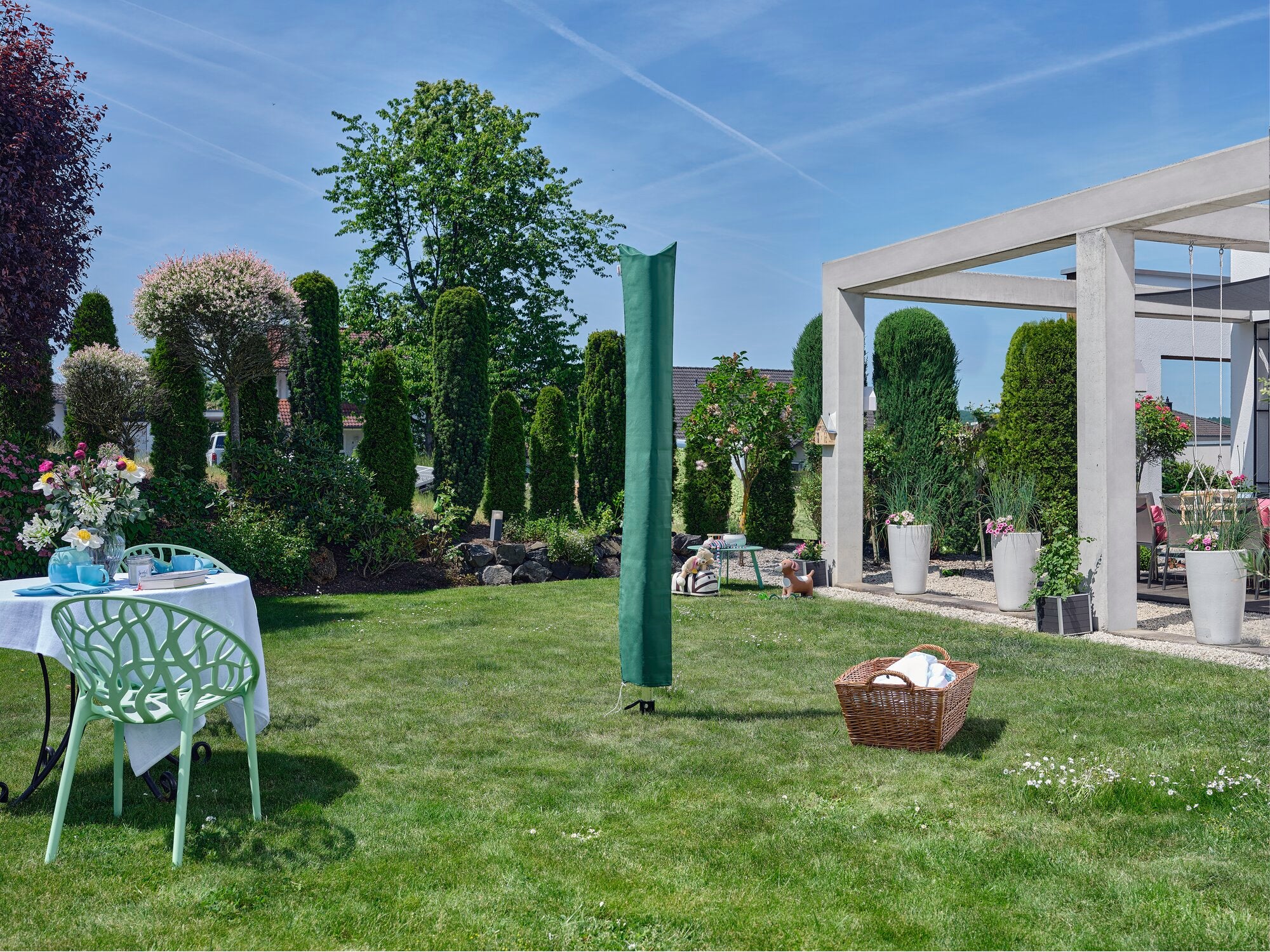Eine grüne Leifheit Linomatic 500 Deluxe Wäschespinne steht in einem gepflegten Garten. Im Hintergrund sind Pflanzen und ein moderner Pavillon zu sehen. Ein Tisch mit Blumen und ein Wäschekorb ergänzen die Szene.