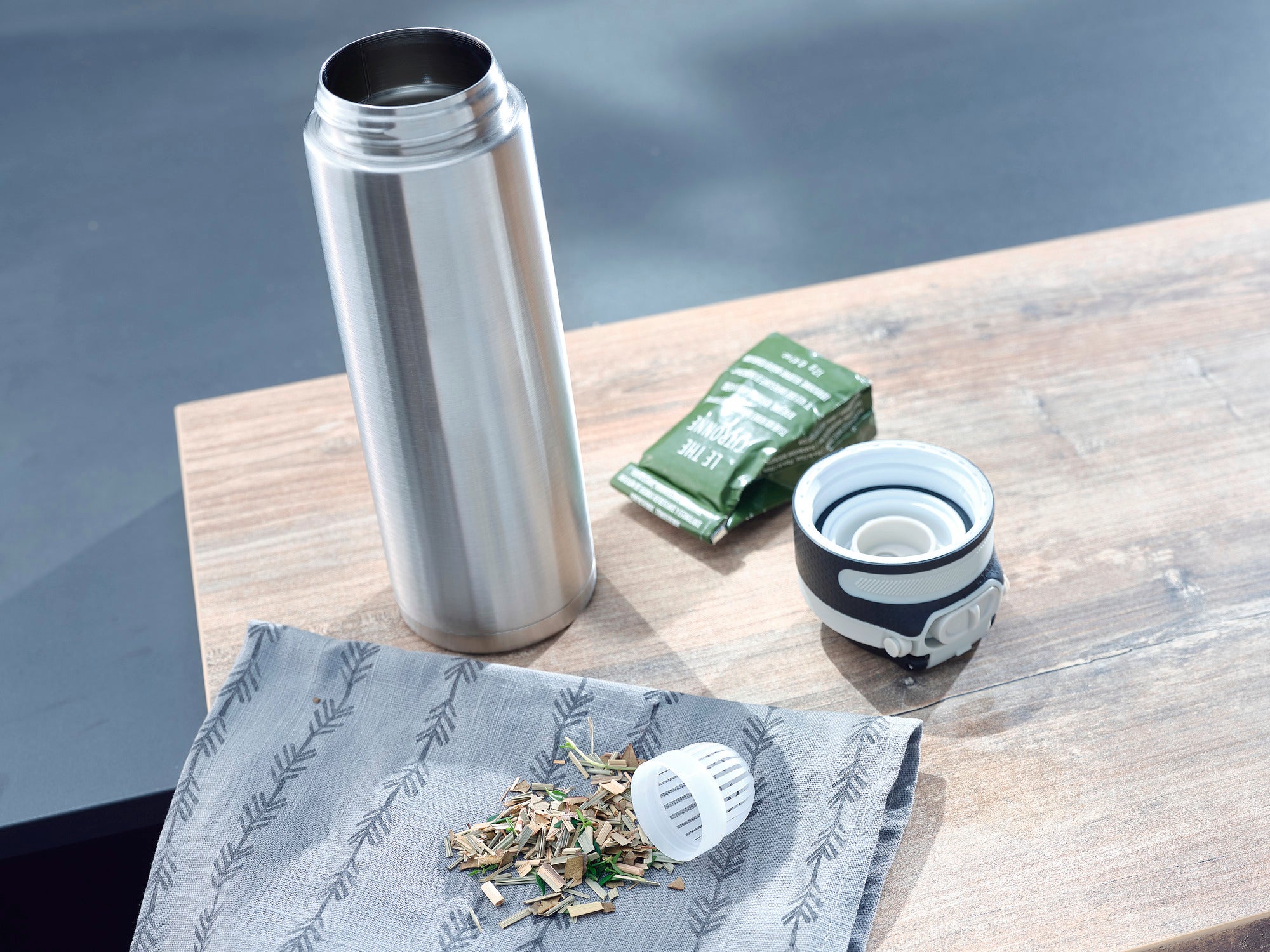 Tasse isotherme Flip 600 ml en argent sur une table en bois. La tasse est ouverte, à côté se trouvent des feuilles de thé, une passoire à thé et un sachet de thé. Idéal pour les déplacements.