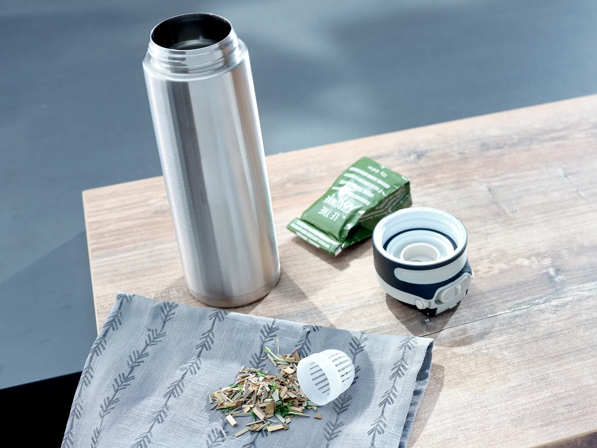 Izolowany kubek Flip 600 ml w kolorze srebrnym na drewnianym stole. Kubek jest otwarty, obok znajdują się liście herbaty, sitko do herbaty i torebka herbaty. Idealny w podróży.