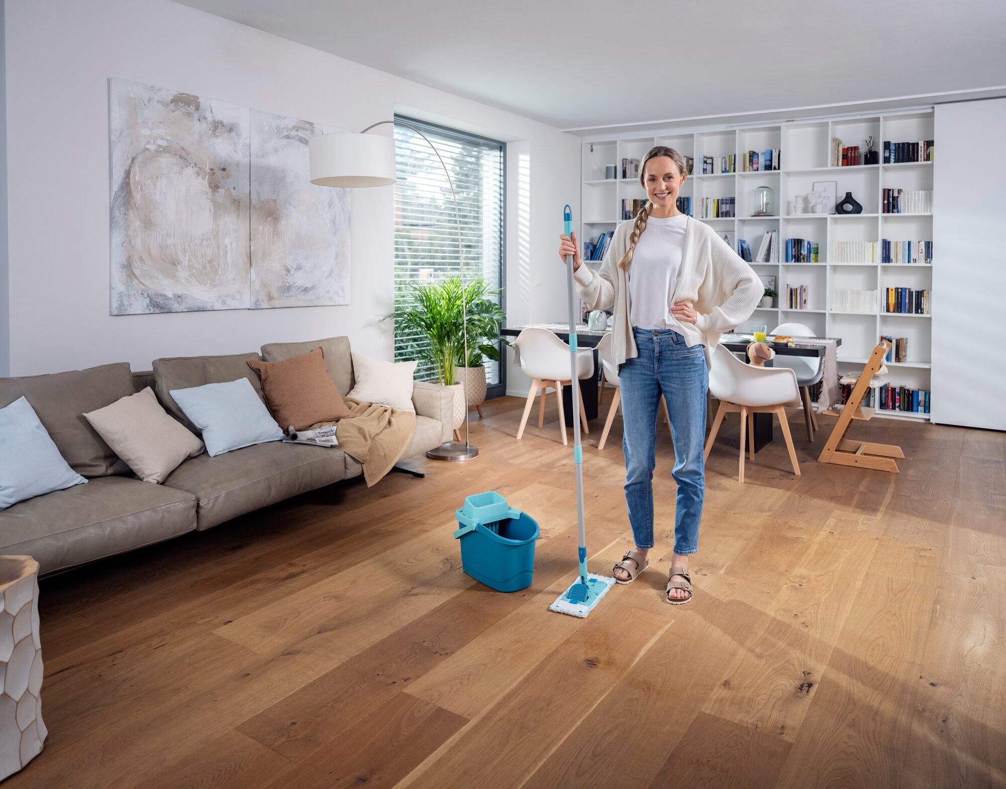 Une femme se tient dans un salon moderne avec une raclette POWER CLEAN. La pièce est lumineuse et élégamment meublée, avec une grande bibliothèque et un canapé confortable.