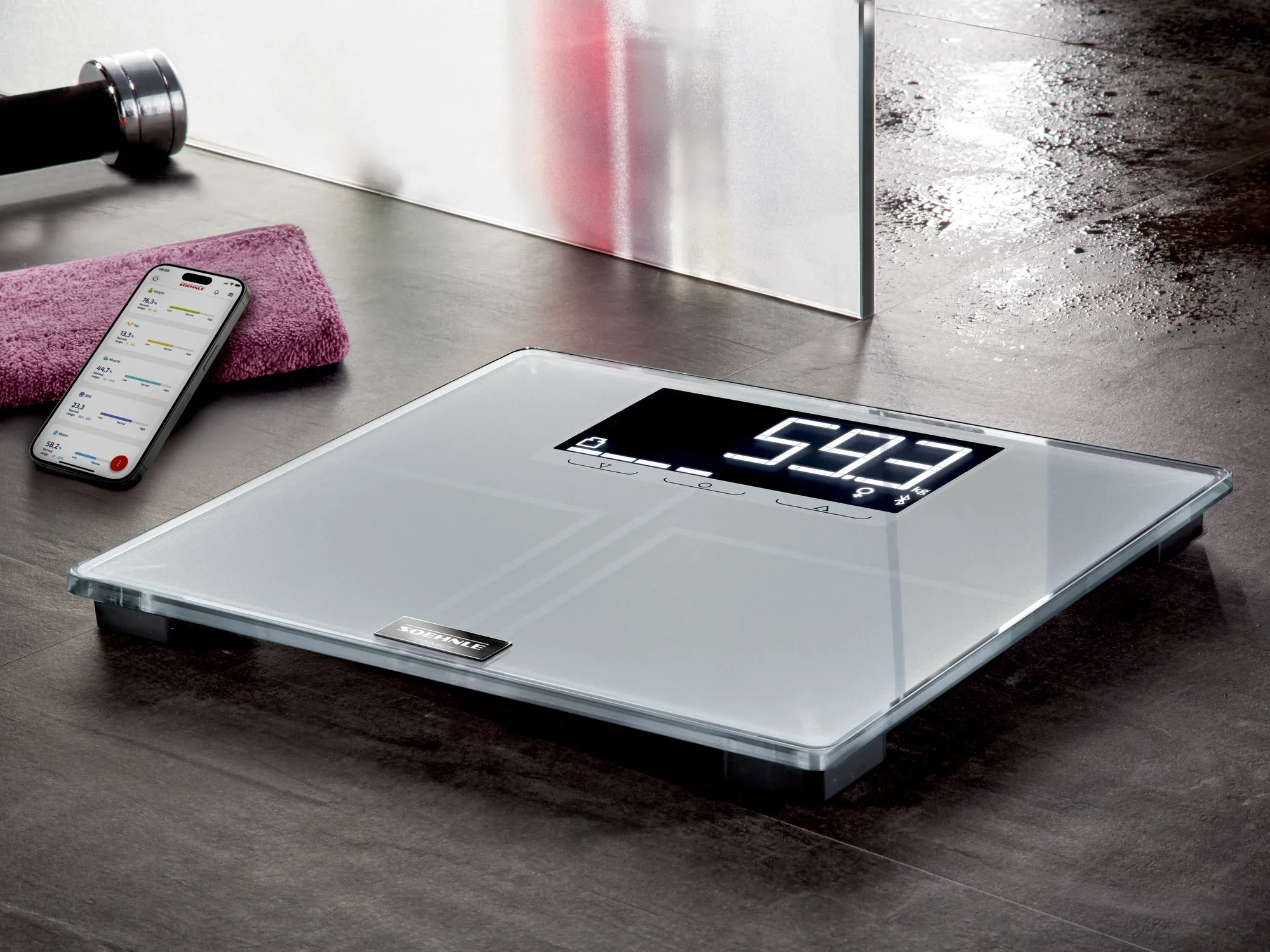 Cyfrowa waga łazienkowa Shape Sense Connect 200 na podłodze w łazience. Waga wyświetla wagę 59,3 kg. Obok znajduje się smartfon z otwartą aplikacją.