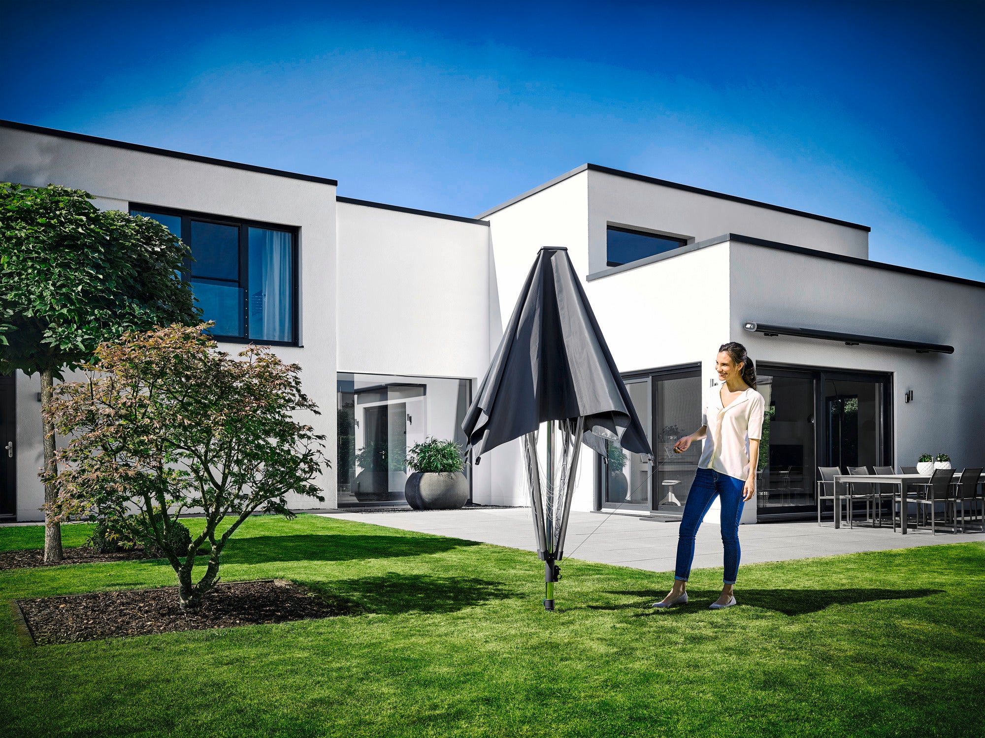 Eine Frau steht im Garten eines modernen Hauses und bedient die Wäschespinne LinoProtect 400. Die Wäschespinne ist zusammengeklappt und bietet Schutz vor Witterungseinflüssen.