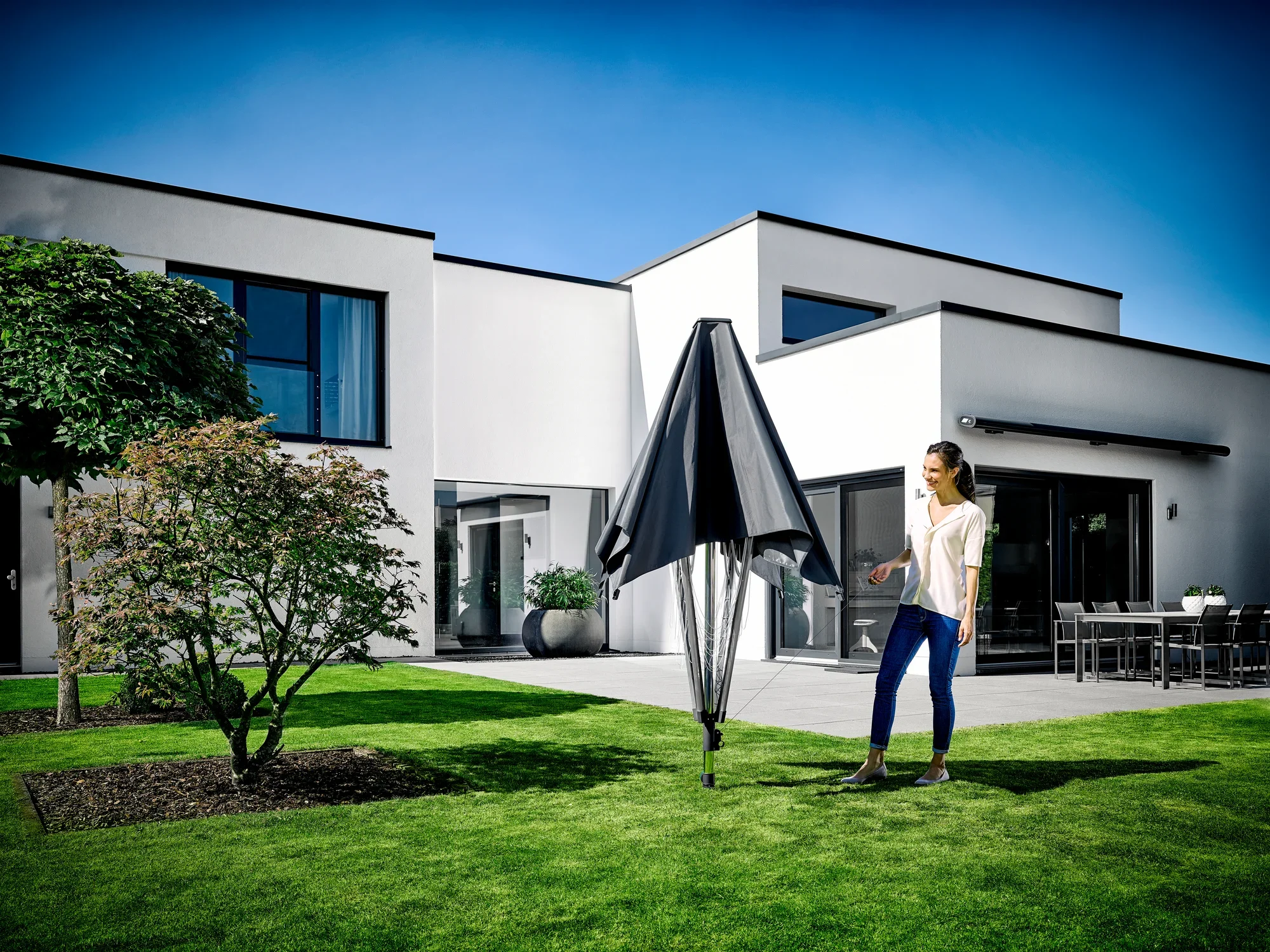 Une femme se tient dans le jardin d'une maison moderne et utilise le séchoir à linge LinoProtect 400. Le séchoir à linge est replié et offre une protection contre les intempéries.