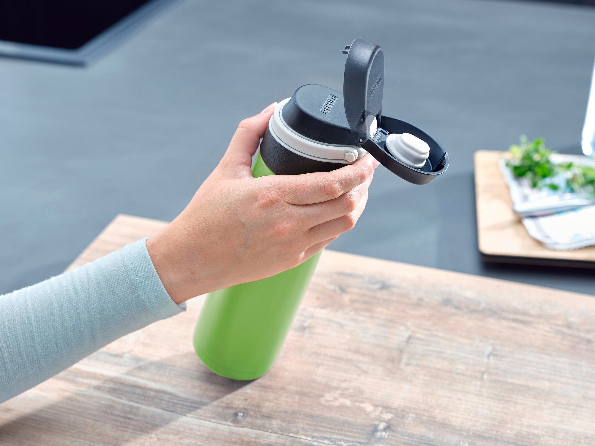 Isolierbecher Flip 600ml in Kiwi-Grün wird auf einem Tisch verwendet. Eine Hand öffnet den Deckel, um den praktischen Verschluss zu zeigen. Ideal für unterwegs.