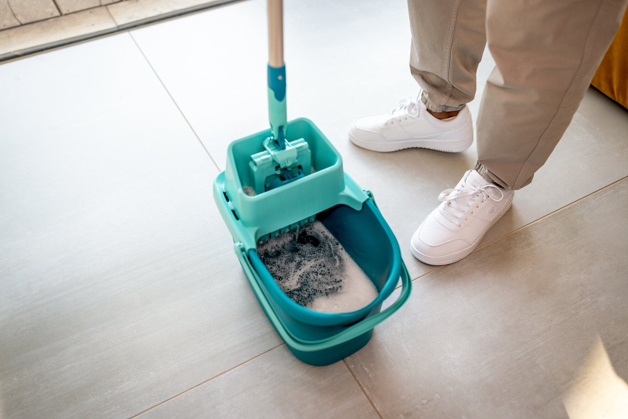 Leifheit POWER CLEAN Lavapavimenti-Eimer mit Auswringmechanik auf hellem Fliesenboden, teilweise mit Wasser und Schaum gefüllt, neben einer Person in heller Hose und weißen Turnschuhen.