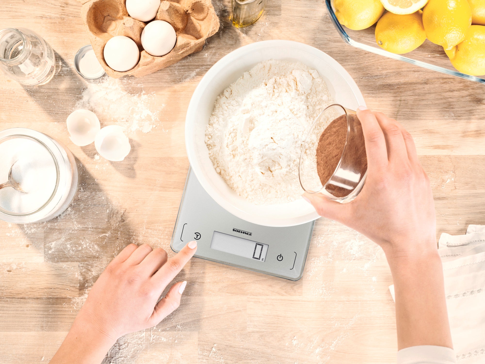 Une balance de cuisine numérique, modèle Page Comfort 300 Slim, est utilisée pour peser de la farine dans un saladier. Une main ajoute le cacao en poudre tandis que l'autre actionne la balance.