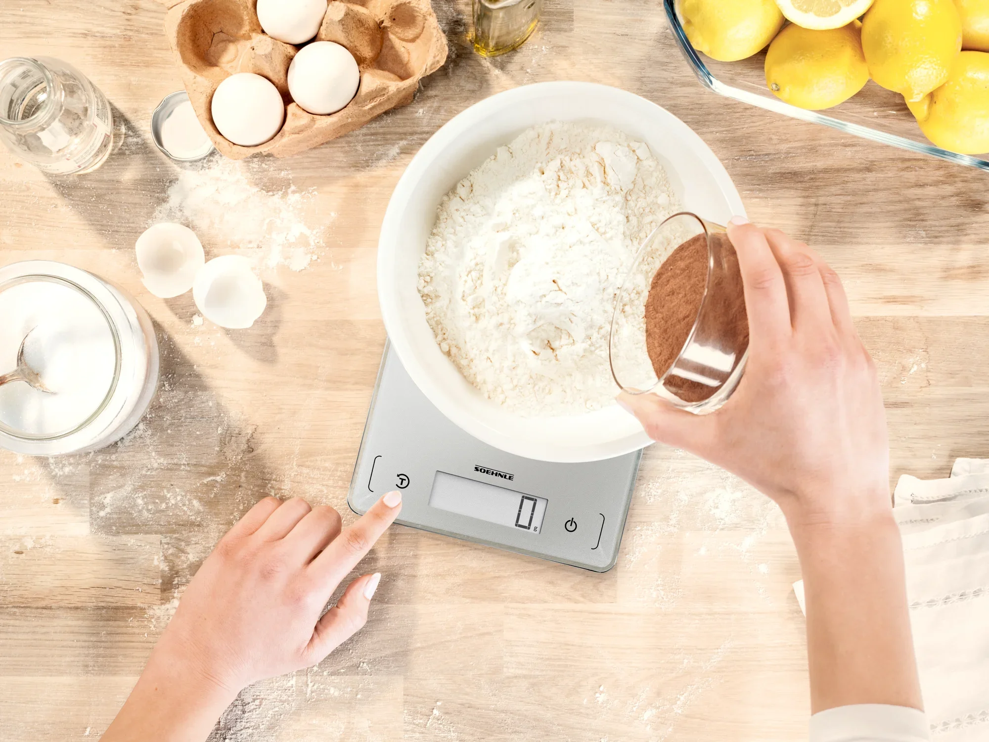 Een digitale keukenweegschaal, model Page Comfort 300 Slim, wordt gebruikt om bloem in een kom af te wegen. Eén hand voegt cacaopoeder toe terwijl de andere de weegschaal bedient.