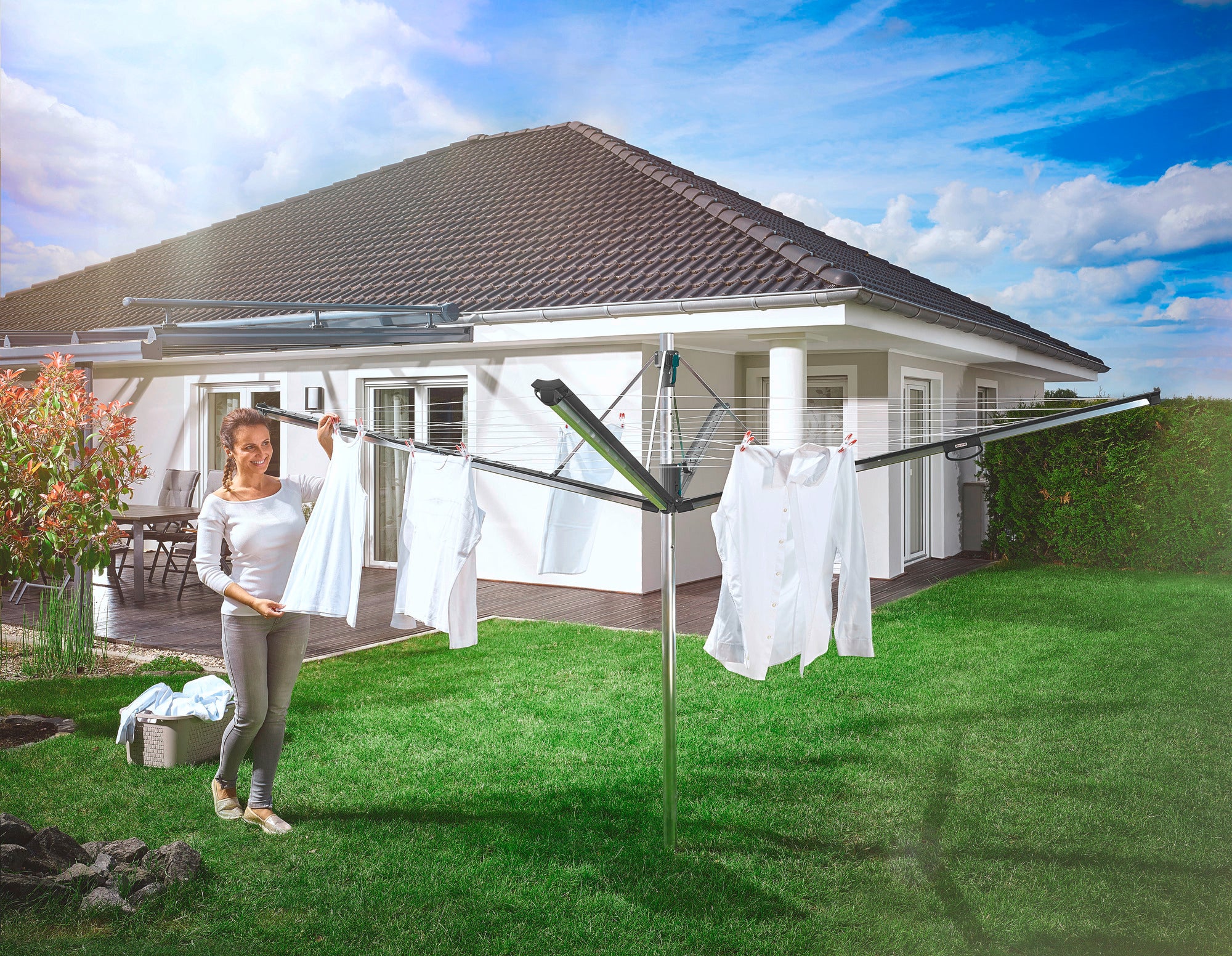 A woman hangs white laundry on a Linomatic 500 Plus rotary clothes dryer in the garden. A modern detached house with a terrace can be seen in the background.