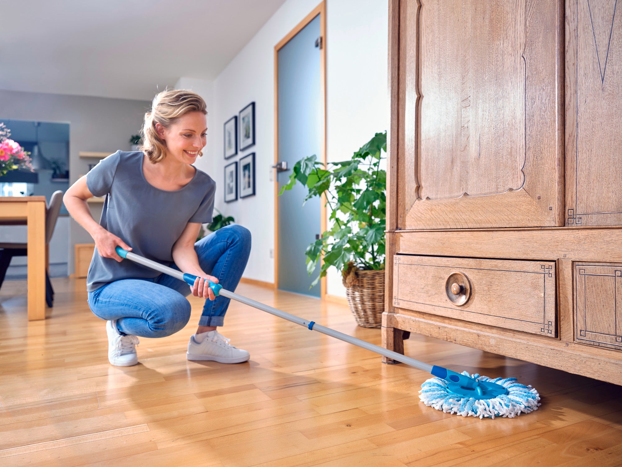 Une femme nettoie le sol en bois avec la serpillière CLEAN TWIST Ergo. Elle sourit en utilisant la serpillière sous une armoire. La pièce est lumineuse et meublée de façon moderne.