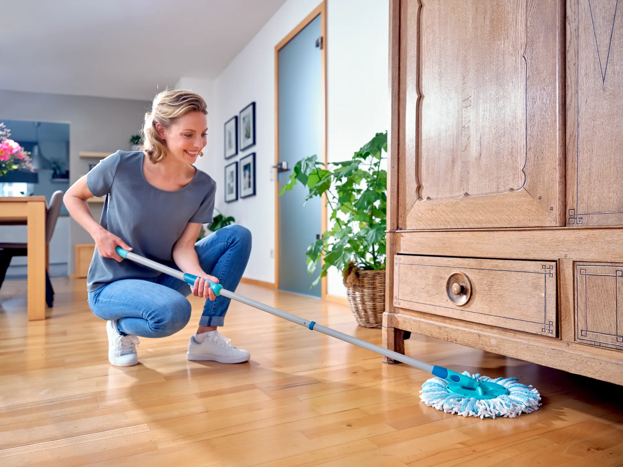 Una mujer limpia el suelo de madera con la mopa CLEAN TWIST Ergo. Sonríe mientras utiliza la mopa debajo de un armario. La habitación es luminosa y tiene una decoración moderna.