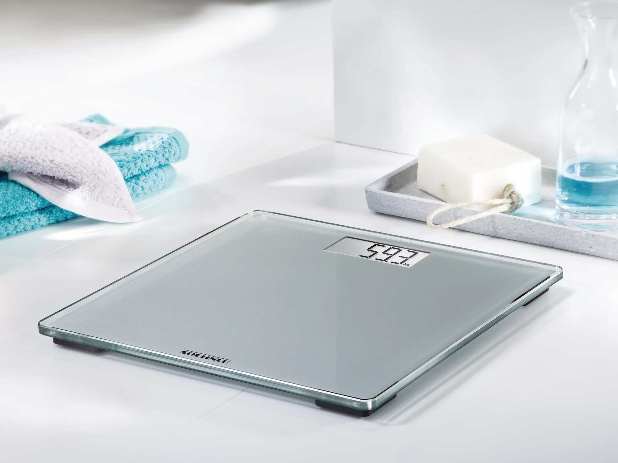 Cyfrowa waga łazienkowa Style Sense Compact 300 stoi na jasnej podłodze w łazience. Wyświetla wagę 54,3 kg. Obok znajdują się ręcznik i mydelniczka.