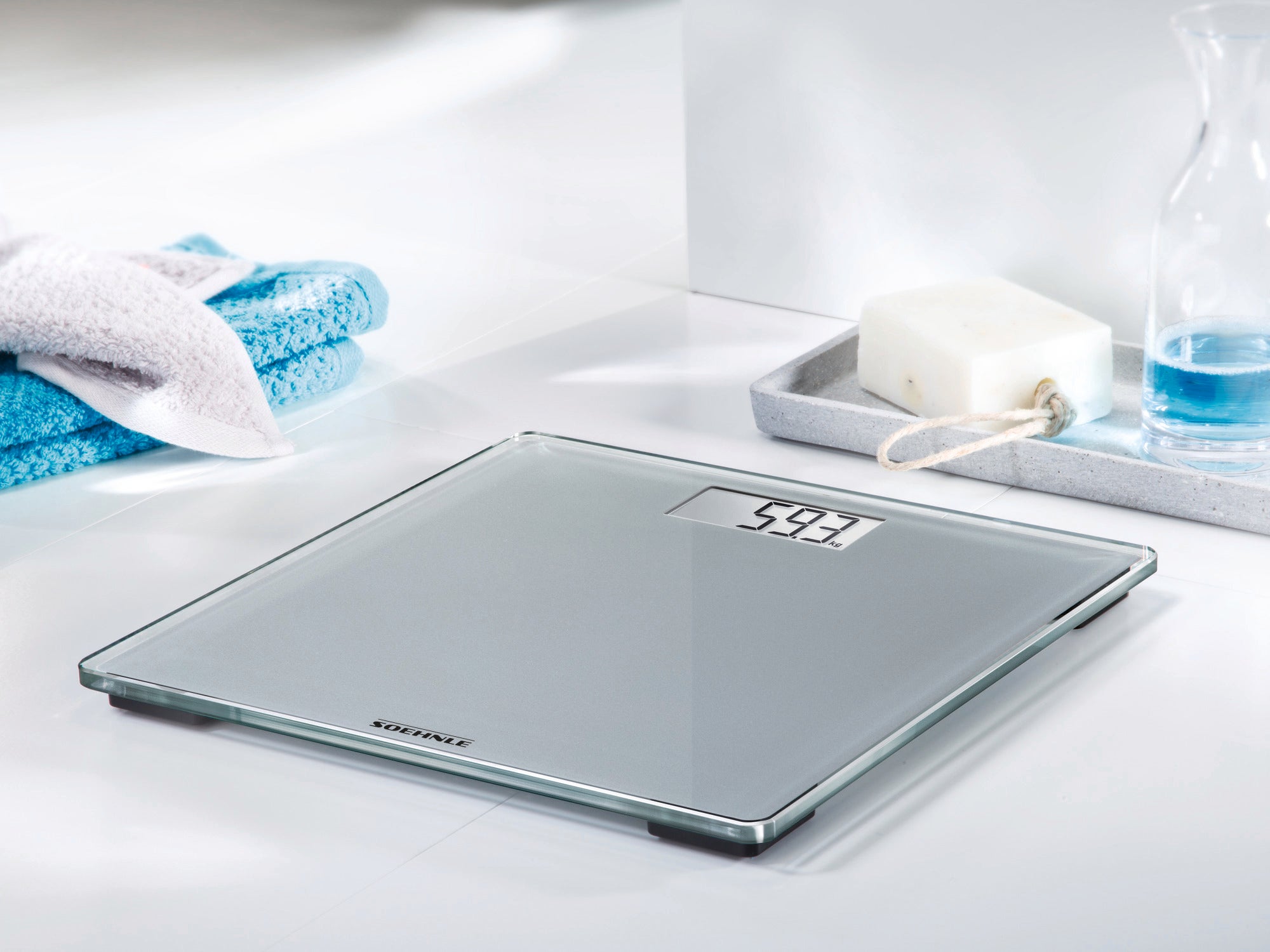 A Style Sense Compact 300 digital bathroom scale stands on a light-coloured bathroom floor. It displays a weight of 54.3 kg. A towel and a soap dish are next to it.
