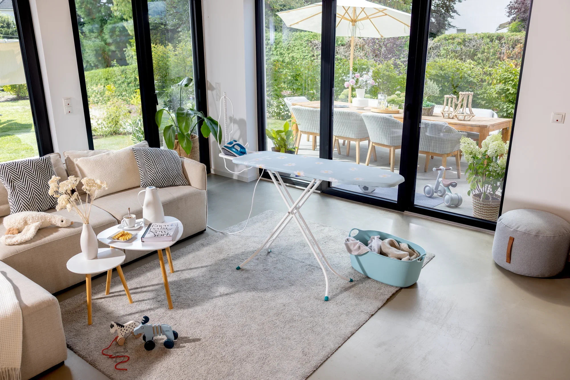 Bügelbrett Leifheit Classic Basic Plus in einem modernen Wohnzimmer mit großen Fenstern. Das Brett steht auf einem Teppich, umgeben von einem Sofa, Tisch und Dekorationen.
