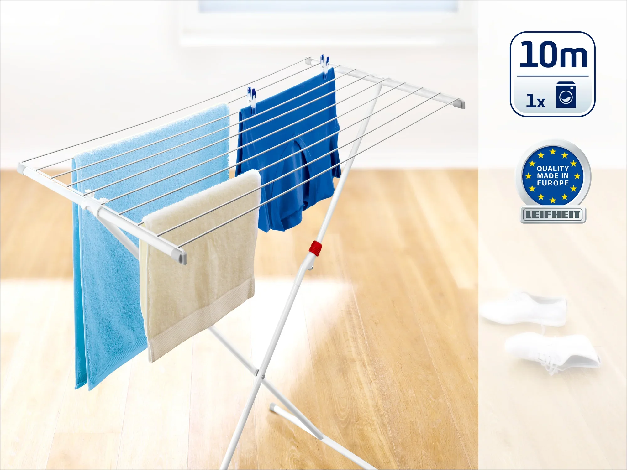 Wolnostojąca suszarka bębnowa z kilkoma liniami, na których wiszą niebieskie i beżowe ręczniki. Suszarka stoi na drewnianej podstawie w jasnym kolorze. Pieczęć wskazuje "Made in Europe".