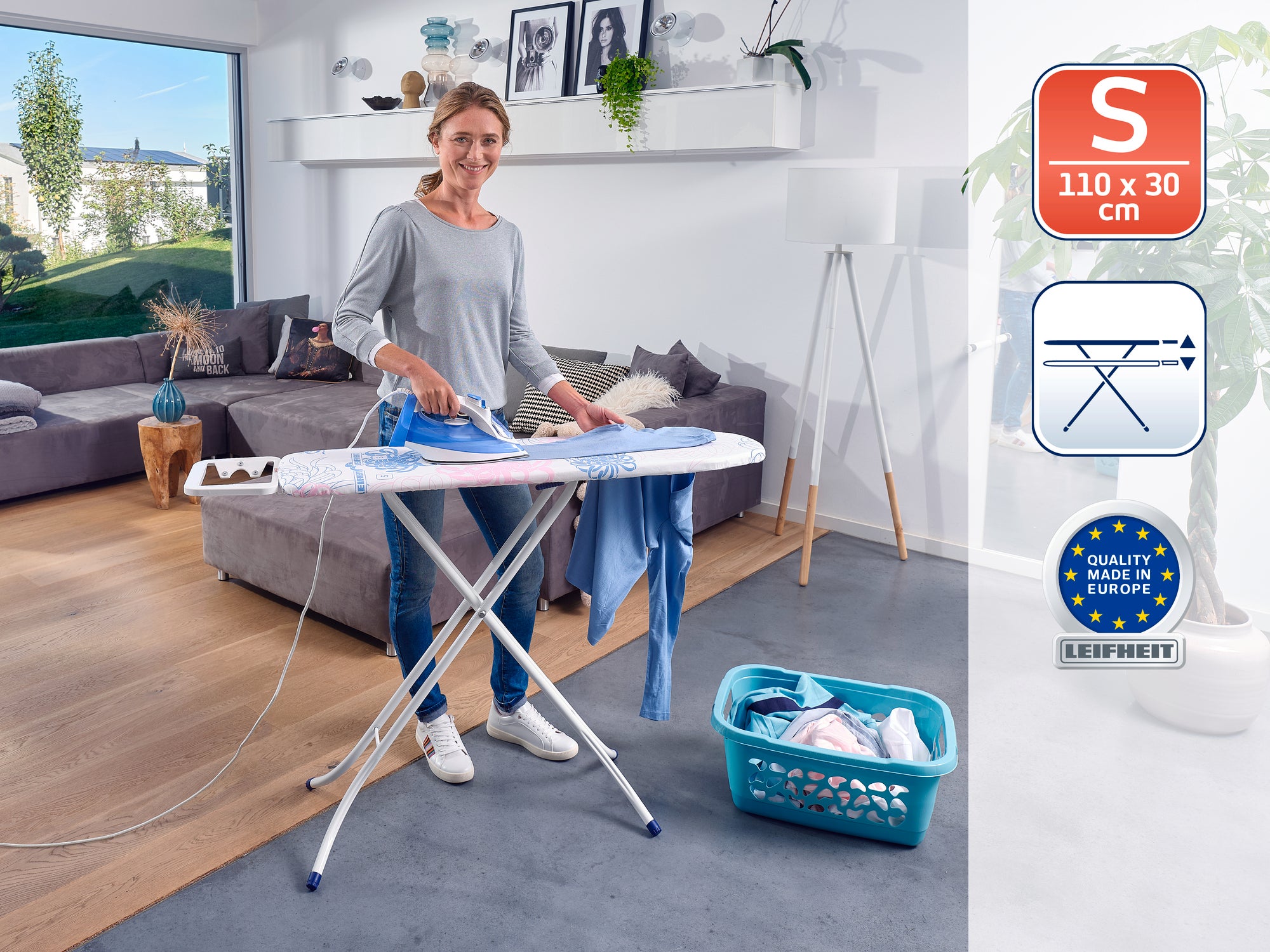 Eine Frau steht in einem modernen Wohnzimmer und bügelt Kleidung auf einem Bügeltisch. Der Tisch ist kompakt, 110 x 30 cm groß, und trägt das Qualitätssiegel „Made in Europe“. Im Hintergrund sind ein Sofa und Pflanzen zu sehen.