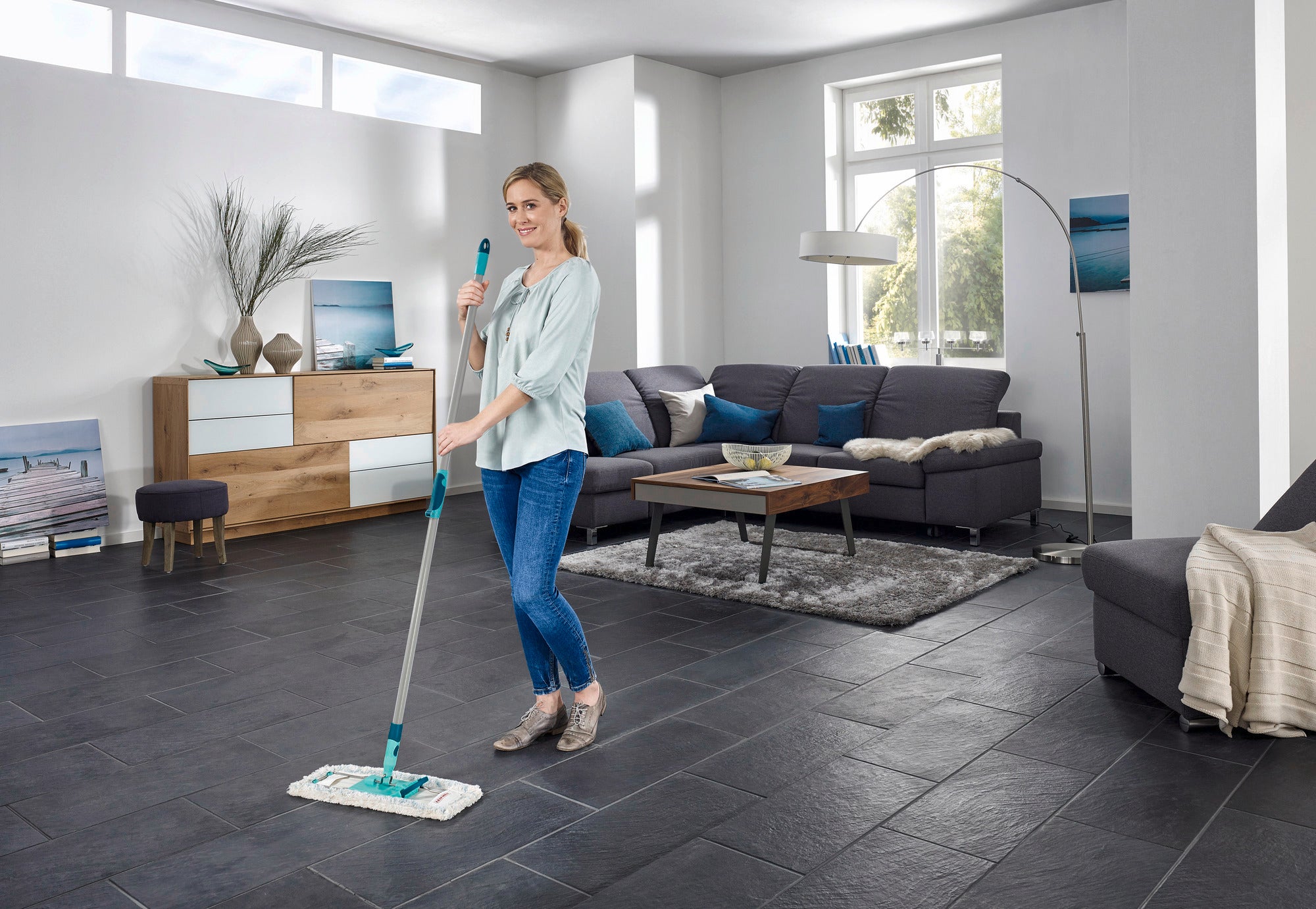 Eine Frau wischt den Boden in einem modernen Wohnzimmer mit dem Bodenwischer Profi XL. Der Raum ist hell und stilvoll eingerichtet, mit einem großen Fenster und eleganten Möbeln.
