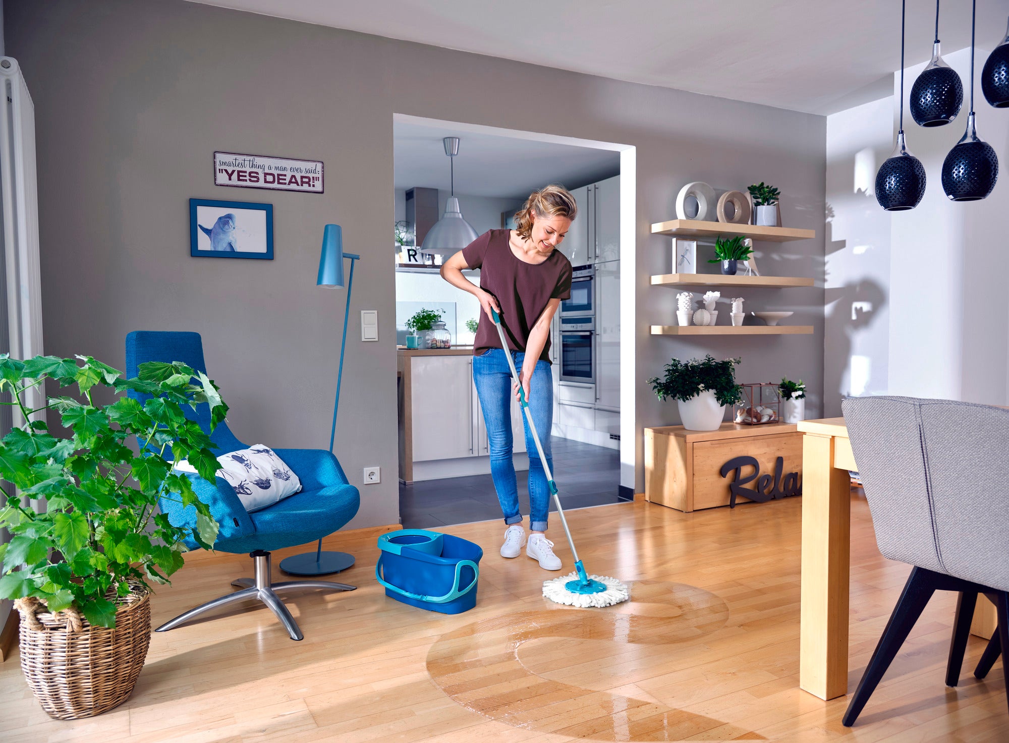 Une femme nettoie le sol en bois d'un salon moderne avec la serpillière Profi XL Rotation Disc. La pièce est lumineuse et élégamment meublée, avec des plantes et des éléments décoratifs.