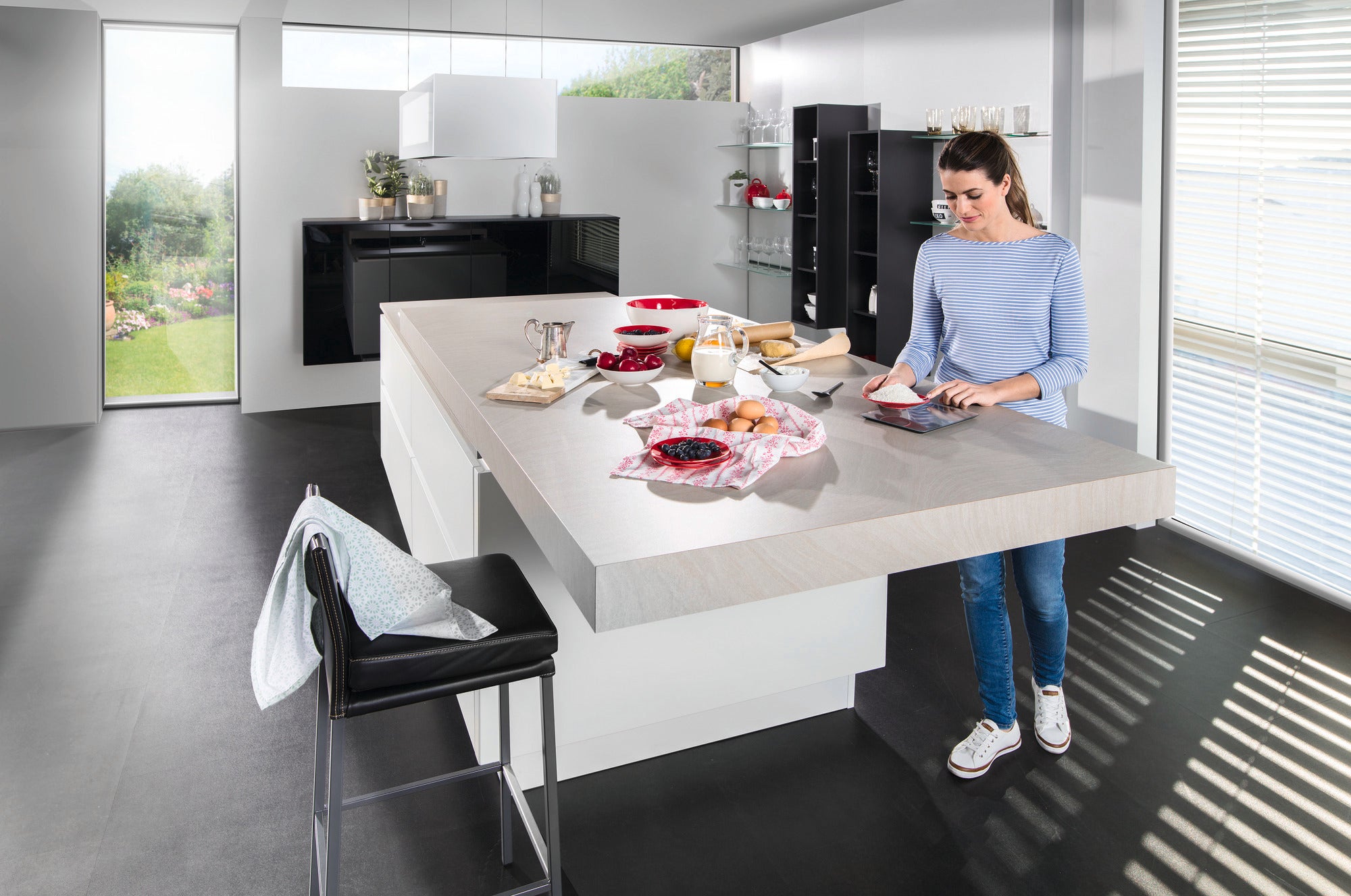 A woman in a modern kitchen uses the Page Comfort 300 Slim digital kitchen scale. Ingredients and a bowl of berries are on the worktop.