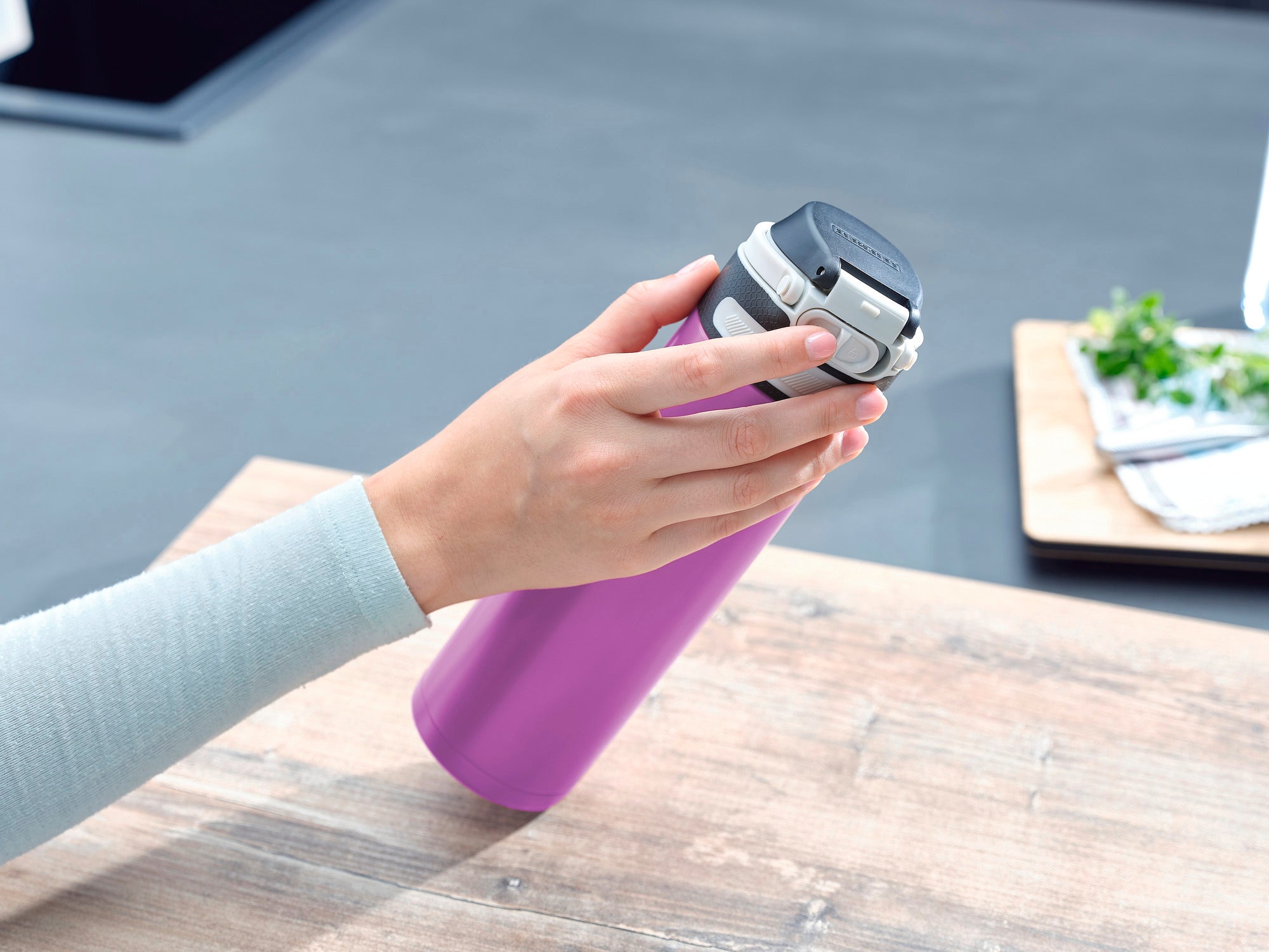 Flip 600 ml insulated mug in purple on a table. A hand opens the lid of the mug. Kitchen utensils and fresh herbs can be seen in the background.