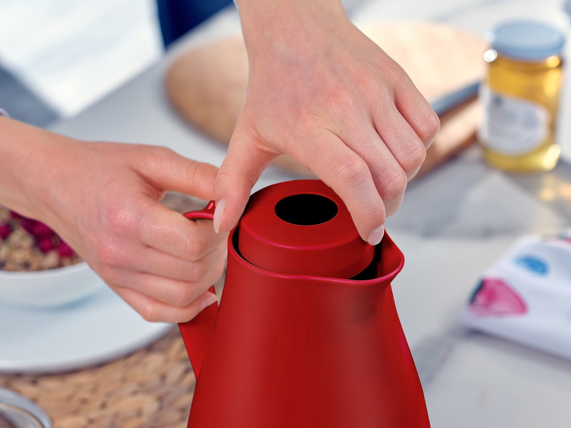 La jarra térmica Harmonic en rojo es abierta por una persona. La jarra está sobre una mesa con utensilios para el desayuno al fondo, entre ellos mermelada y cereales.