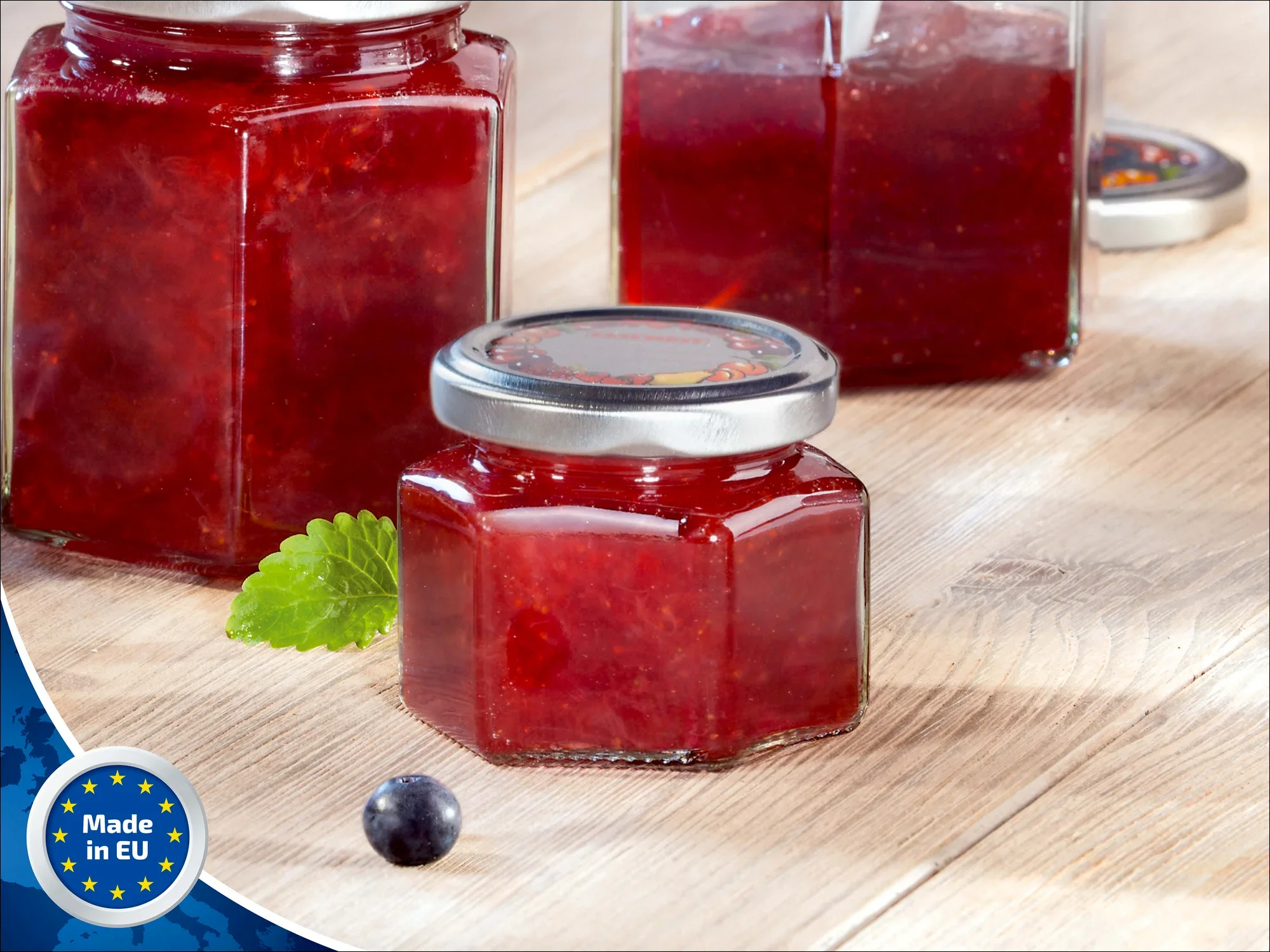 Ein Sechskantglas mit 106 ml Fassungsvermögen, gefüllt mit roter Marmelade, steht auf einem Holztisch. Das Glas hat einen silbernen Deckel. Im Hintergrund sind weitere Gläser sichtbar. Ein "Made in EU"-Logo ist in der Ecke zu sehen.